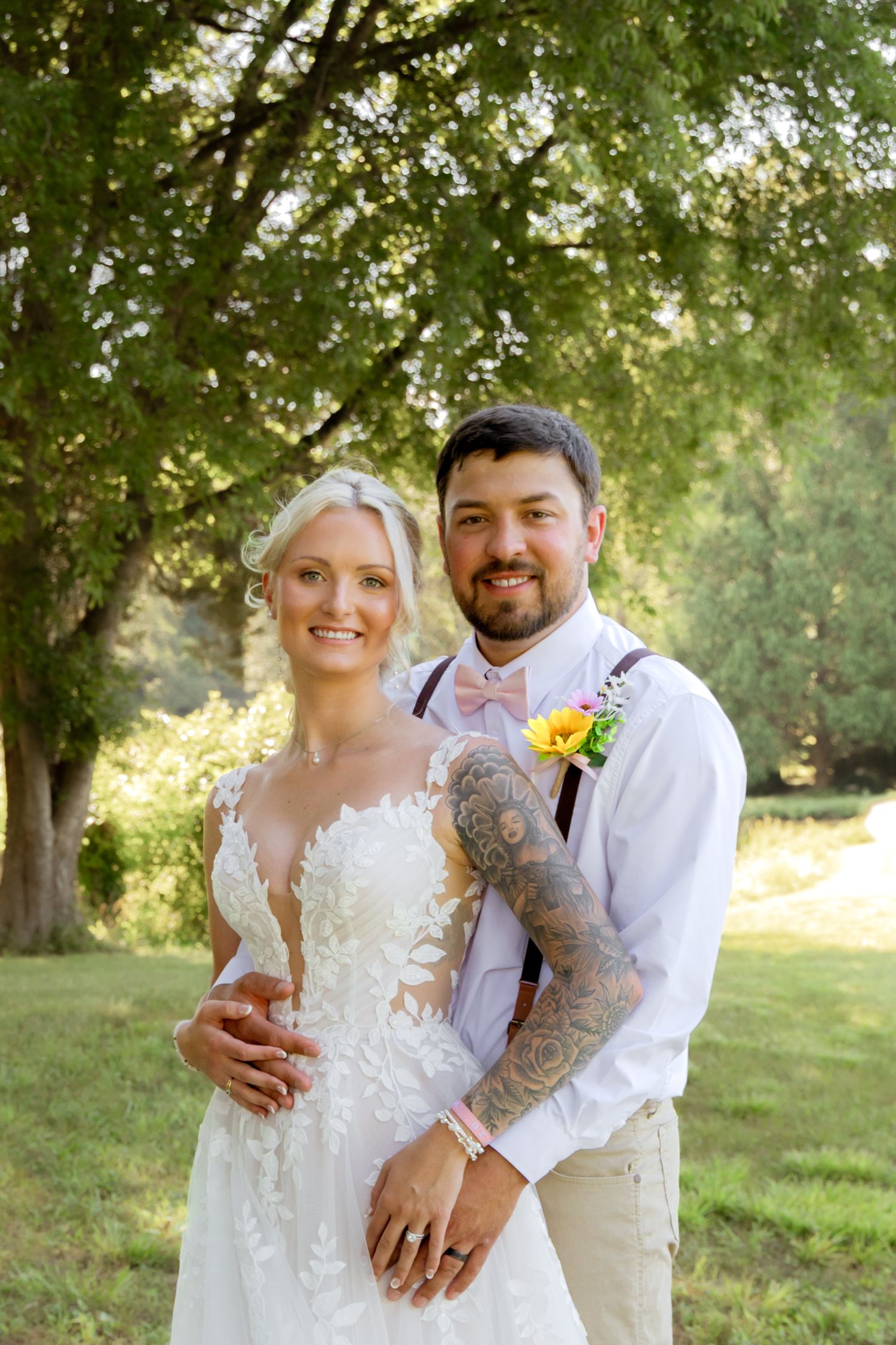 Bride and groom pose outdoors under green trees, groom with tattoos and boutonniere embracing bride in lace wedding dress.