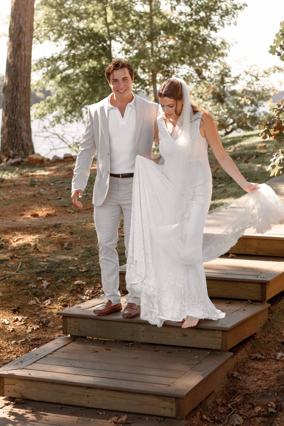 A couple in wedding attire walking down wooden steps in a wooded setting.