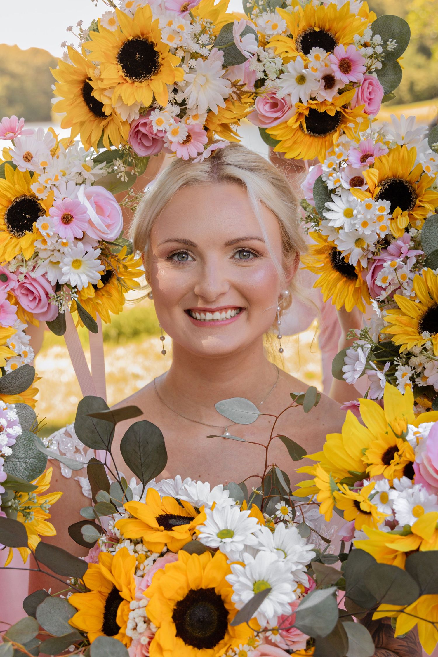 Smiling blonde bride framed by a floral arch of sunflowers, pink roses, and white daisies at an outdoor wedding.