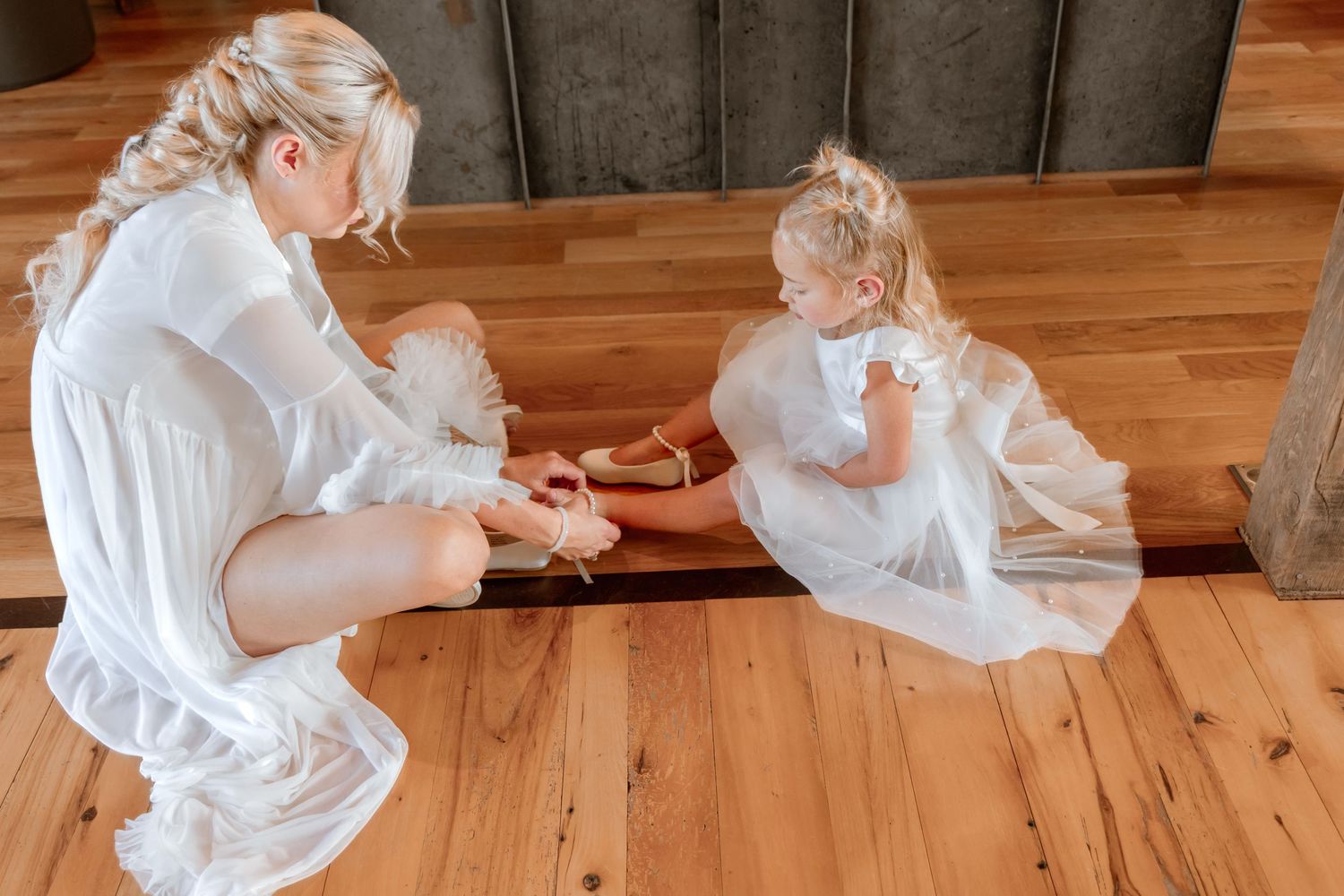 Woman in white dress helping young girl with shoes on wooden floor.
