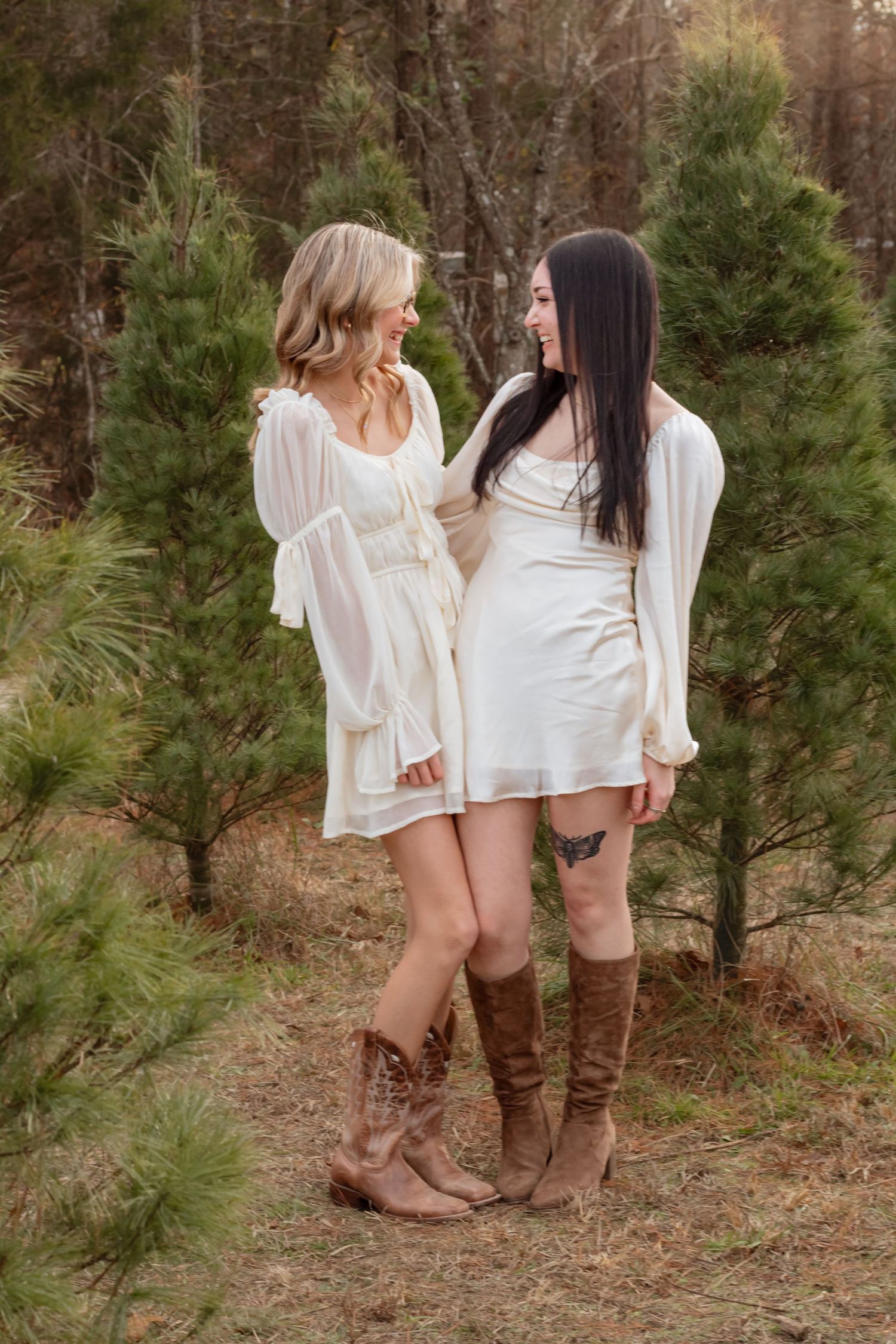 Two women in white dresses and brown boots share a moment among pine trees.