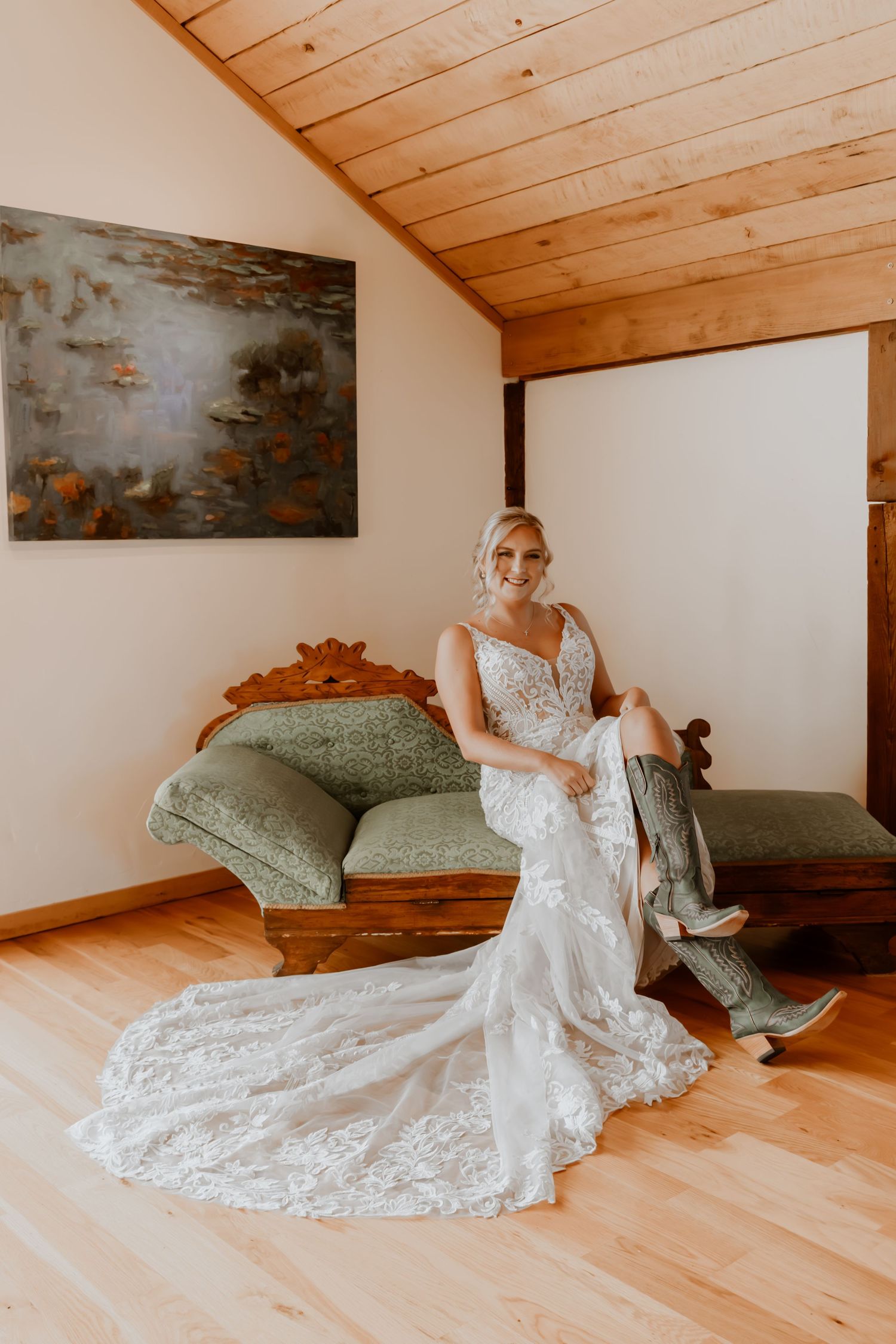Bride in lace wedding gown poses on vintage chaise lounge in rustic room with wood ceiling.