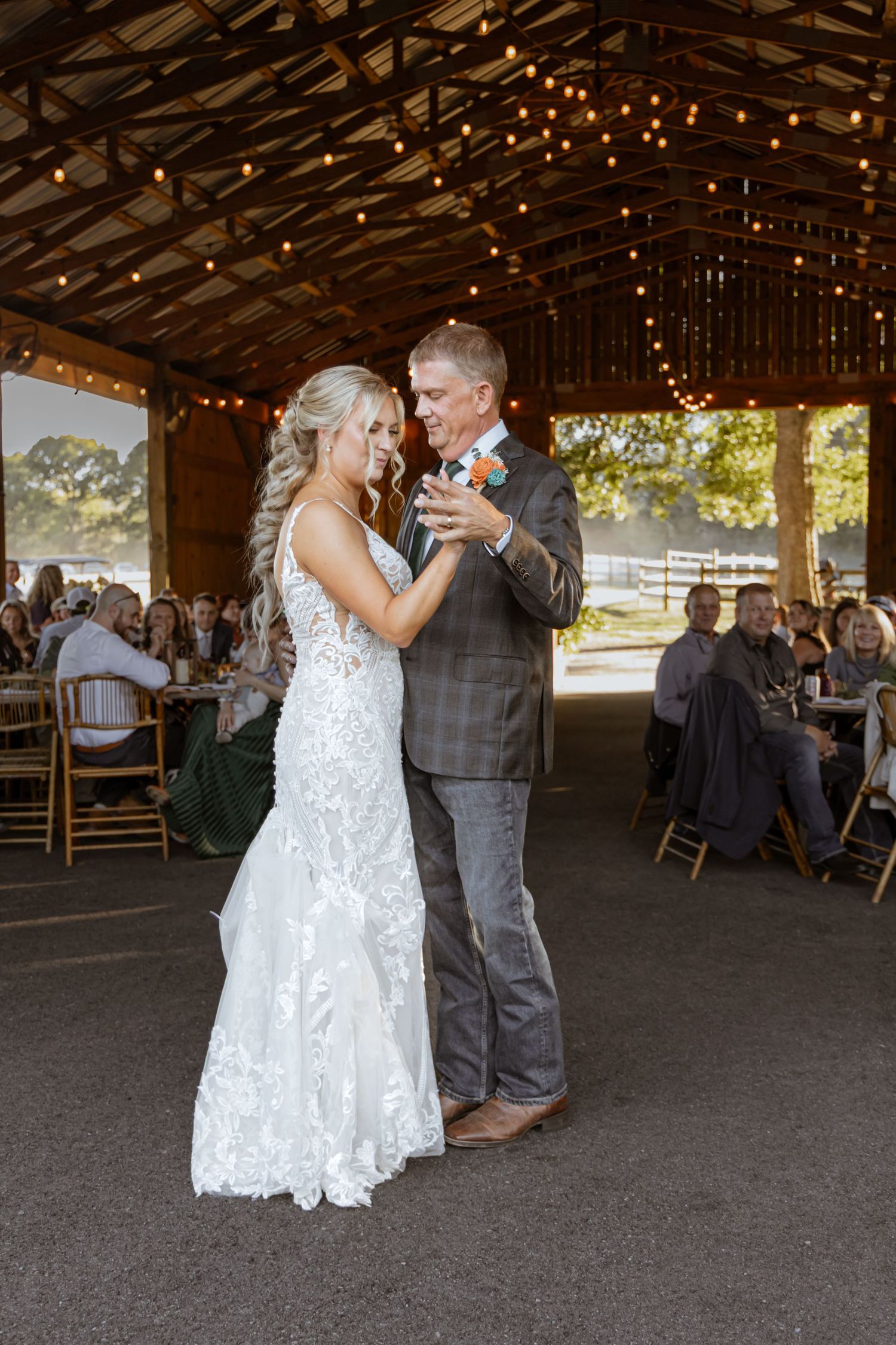 Bride and groom share their first dance under string lights in a rustic barn wedding venue with guests seated nearby.