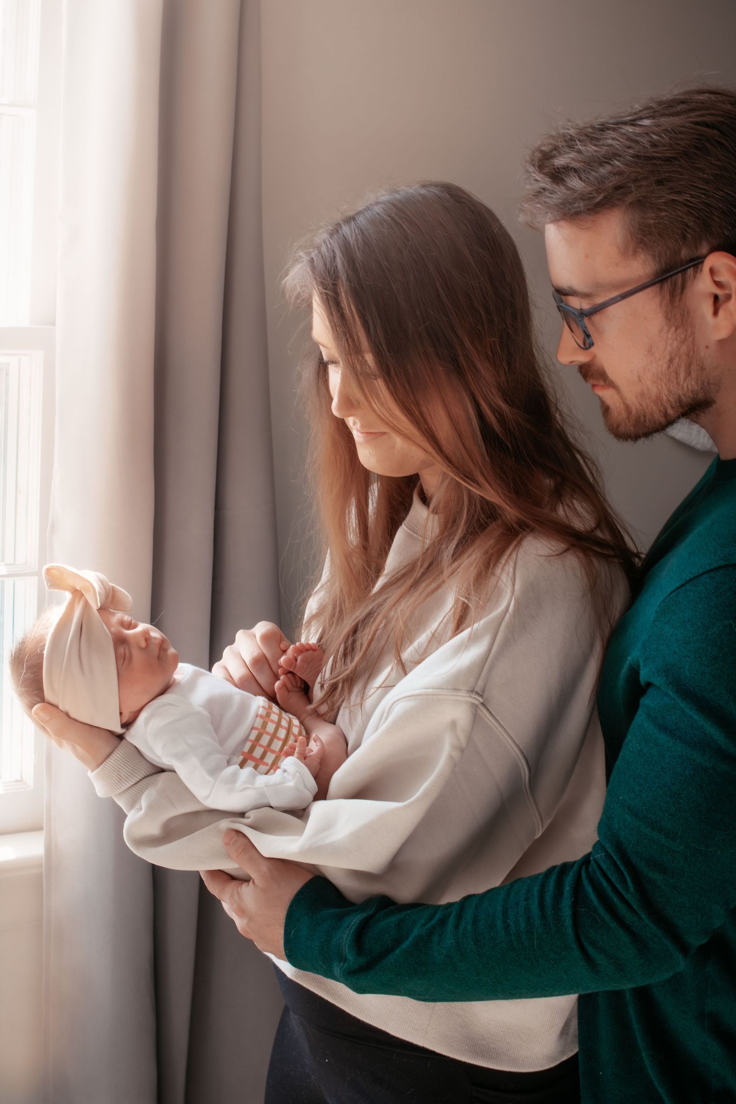 A tender indoor family moment with a newborn baby being held near a window with soft natural light.