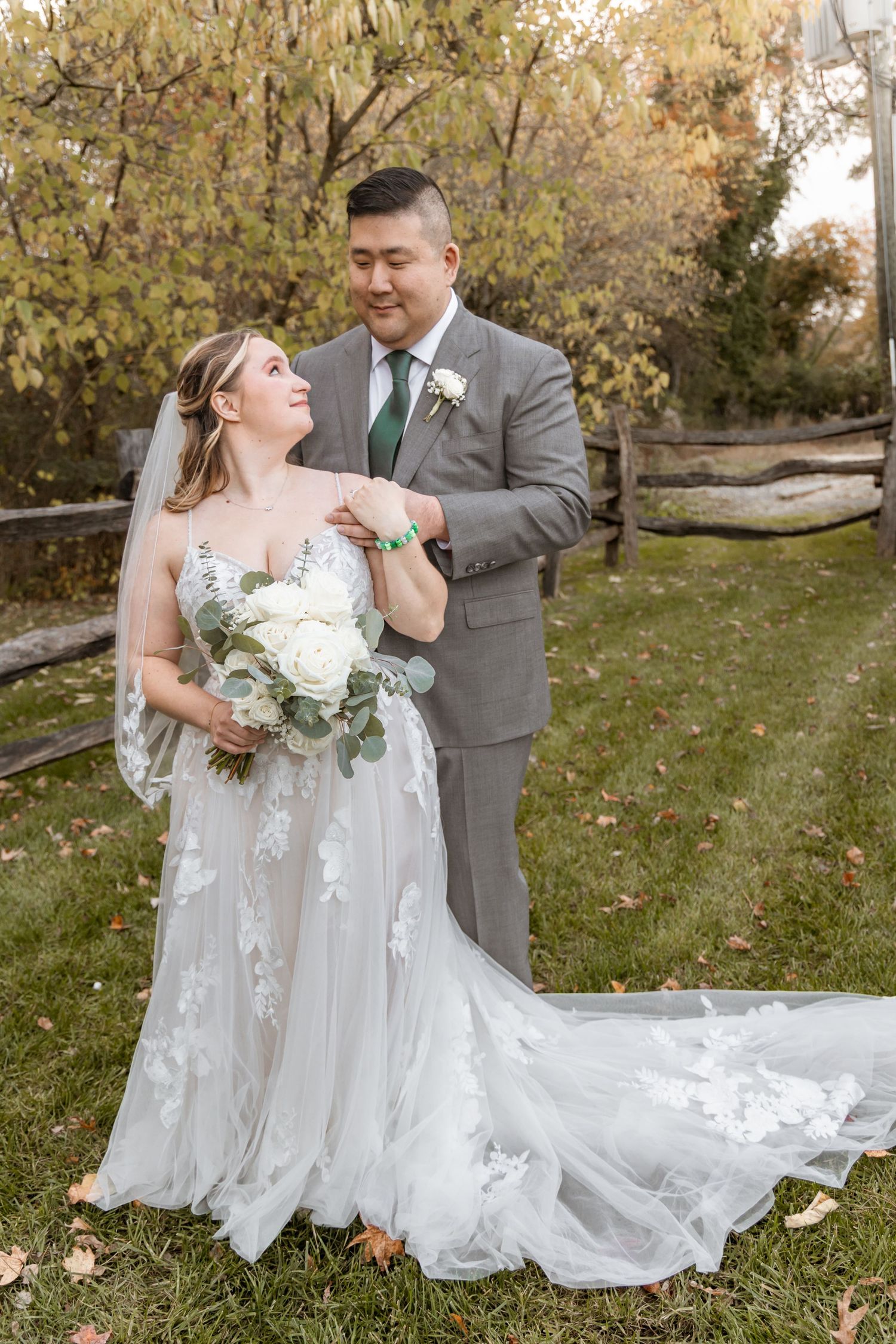 Newlyweds pose together on autumn lawn with bride's flowing white dress trailing behind.