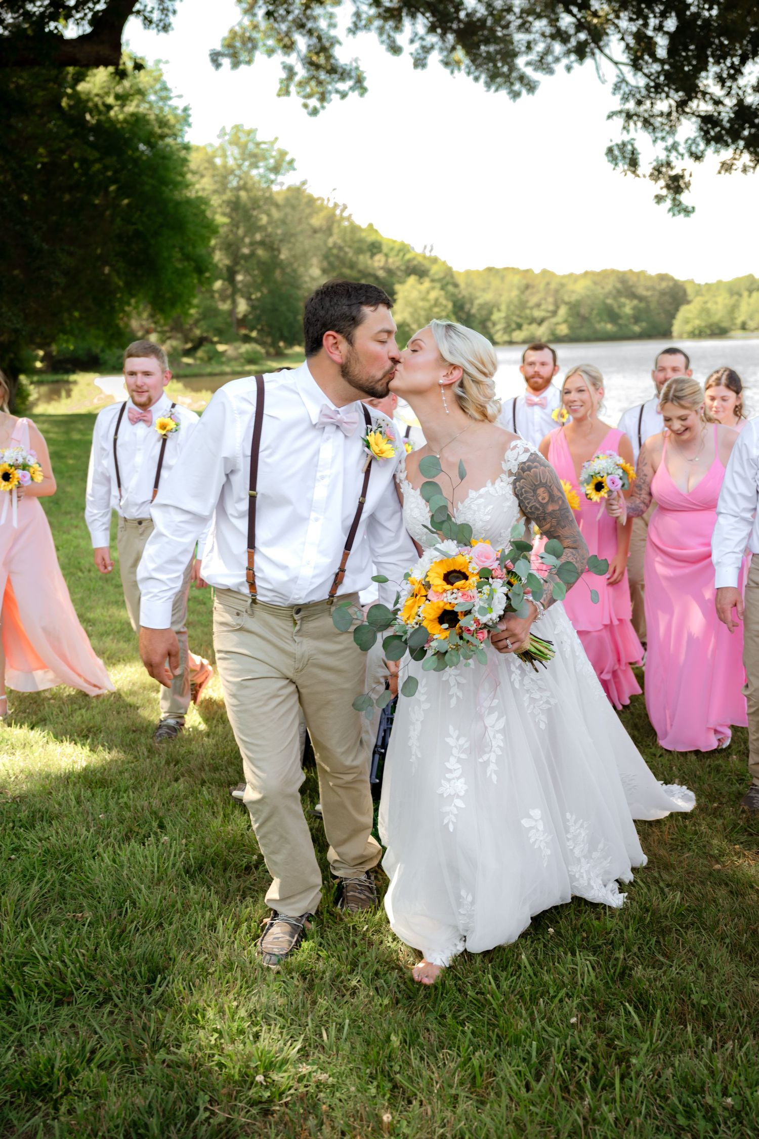 Bride and groom share a kiss outdoors while wedding party in pink and white follows nearby.