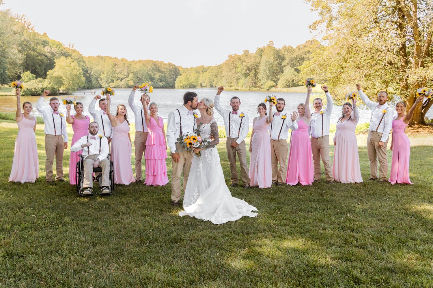 Wedding party celebrating outdoors by a lake, bridesmaids in pink dresses and groomsmen in khaki with suspenders.