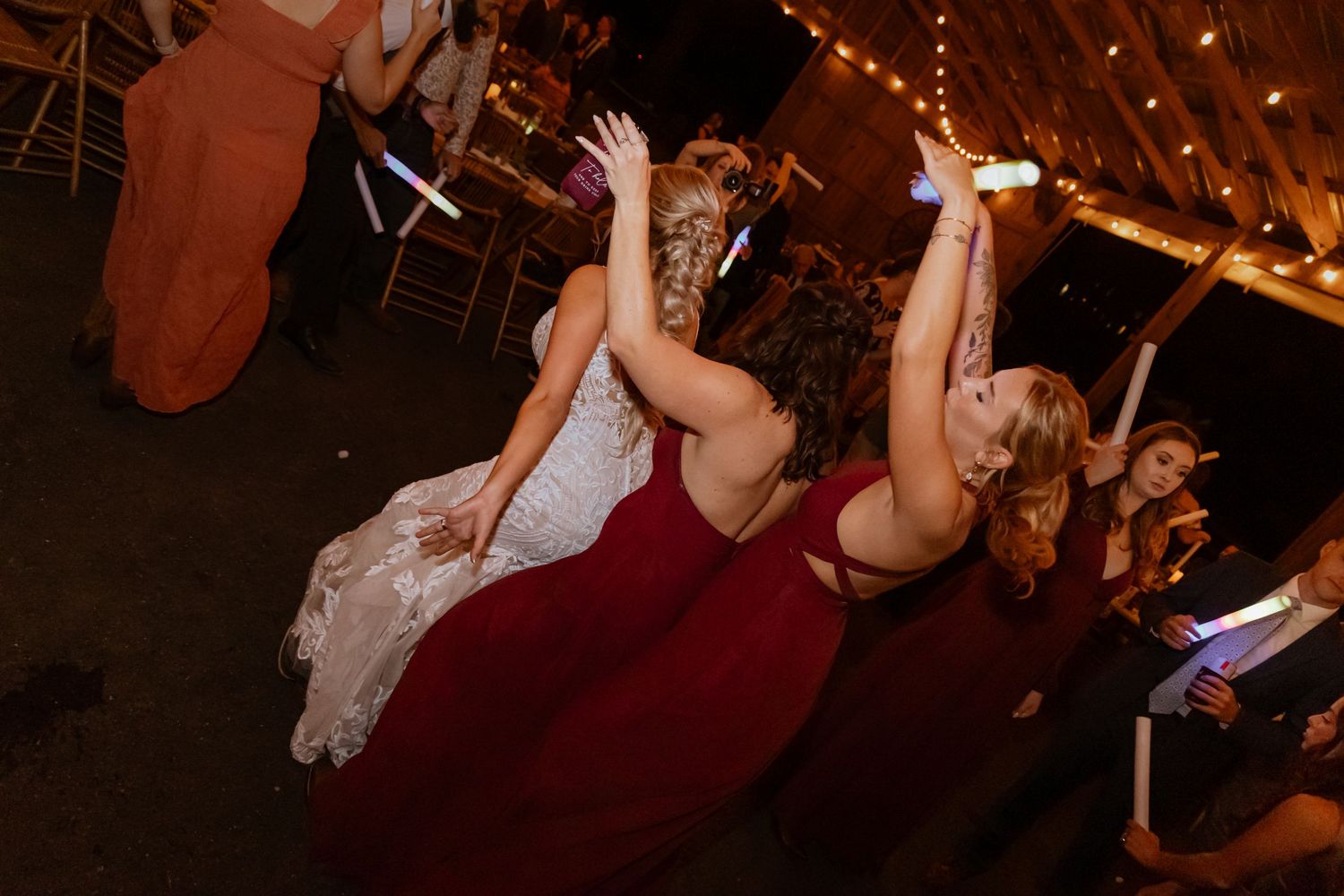 Bride and bridesmaid in red dress dancing joyfully at a wedding reception with string lights overhead.