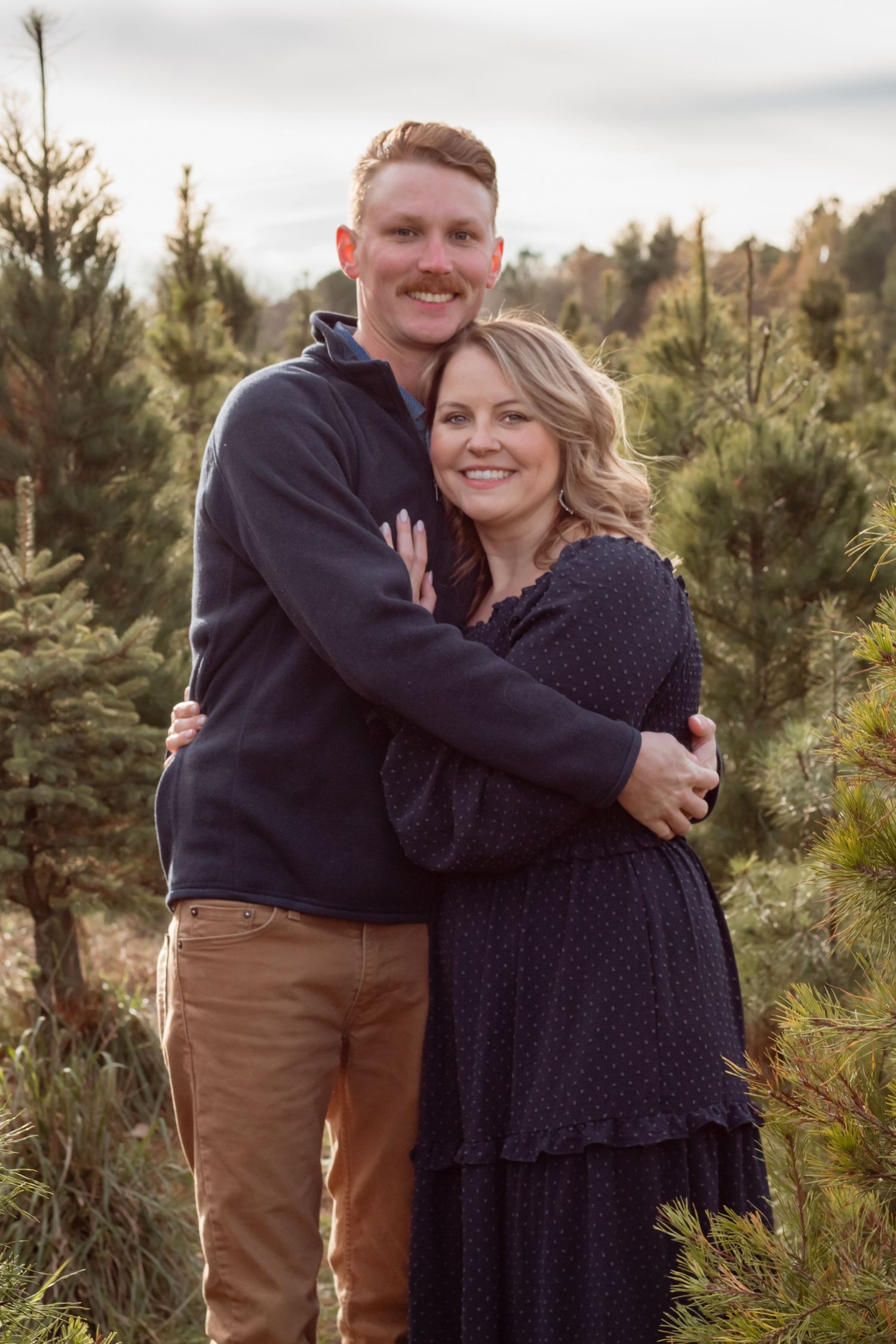 Couple embracing in navy outfits during a romantic photo session among pine trees.