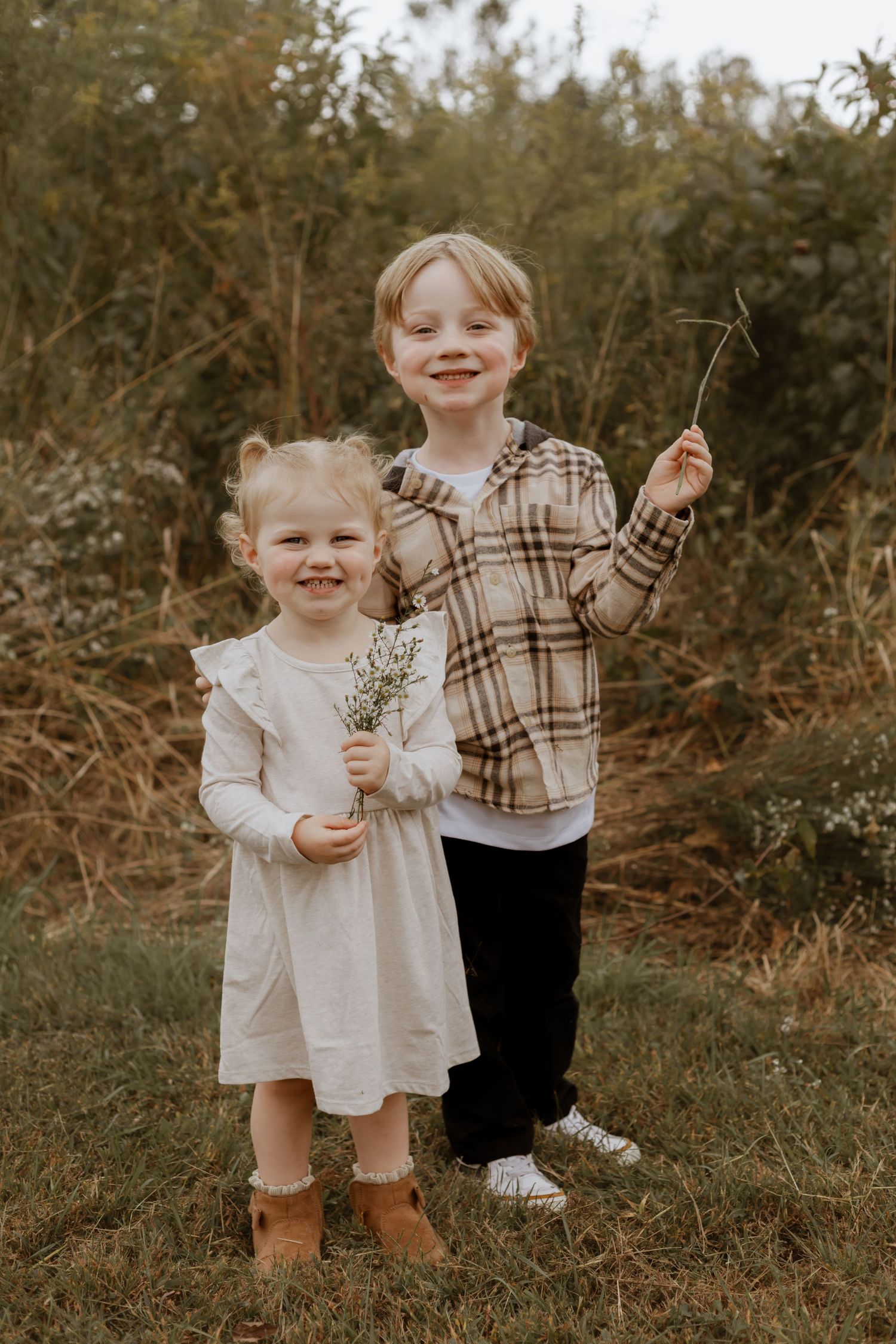 Two children standing in a field wearing white and plaid outfits while holding wildflowers.