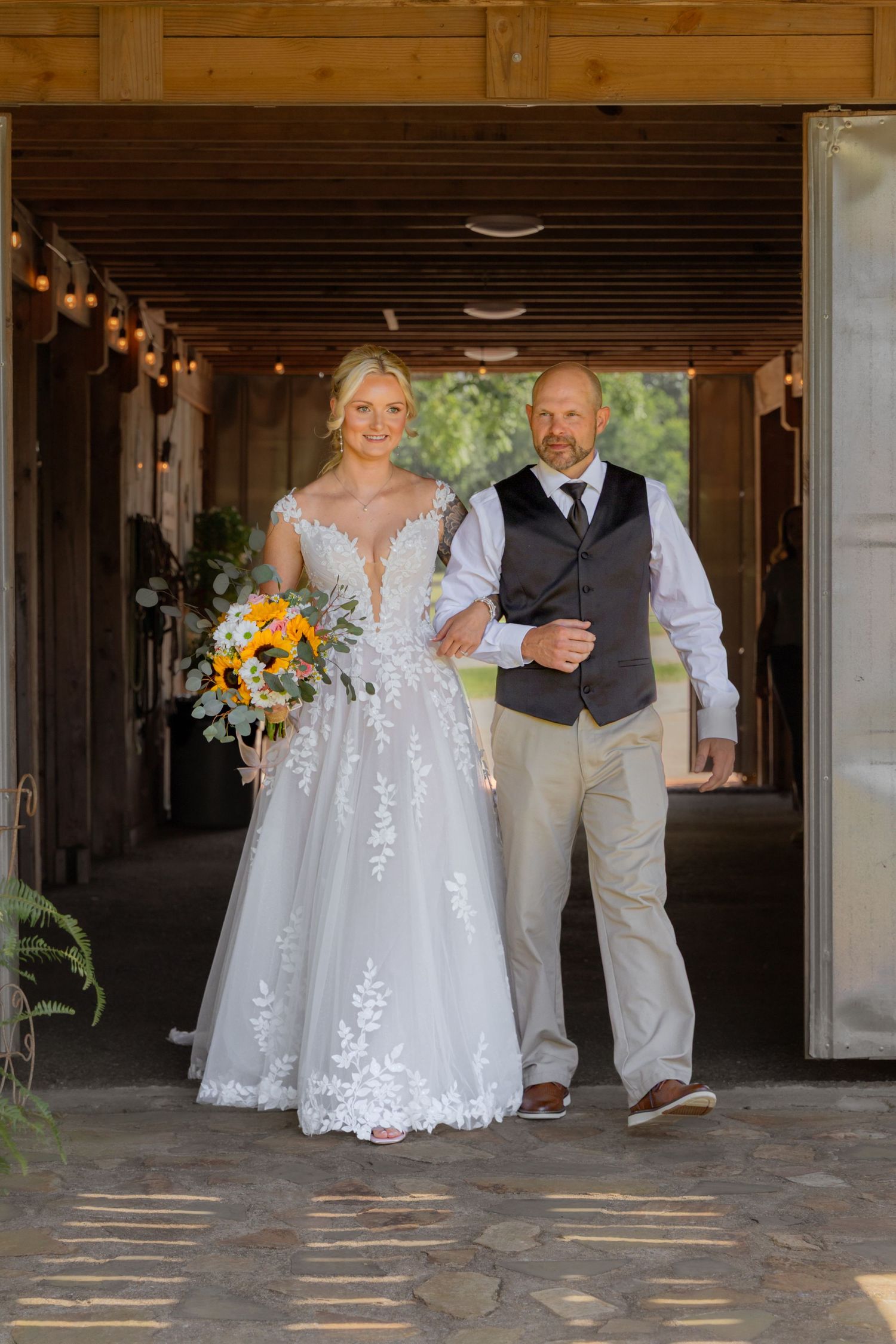 Bride in lace gown holds sunflower bouquet while walking arm-in-arm with escort through rustic barn doorway.