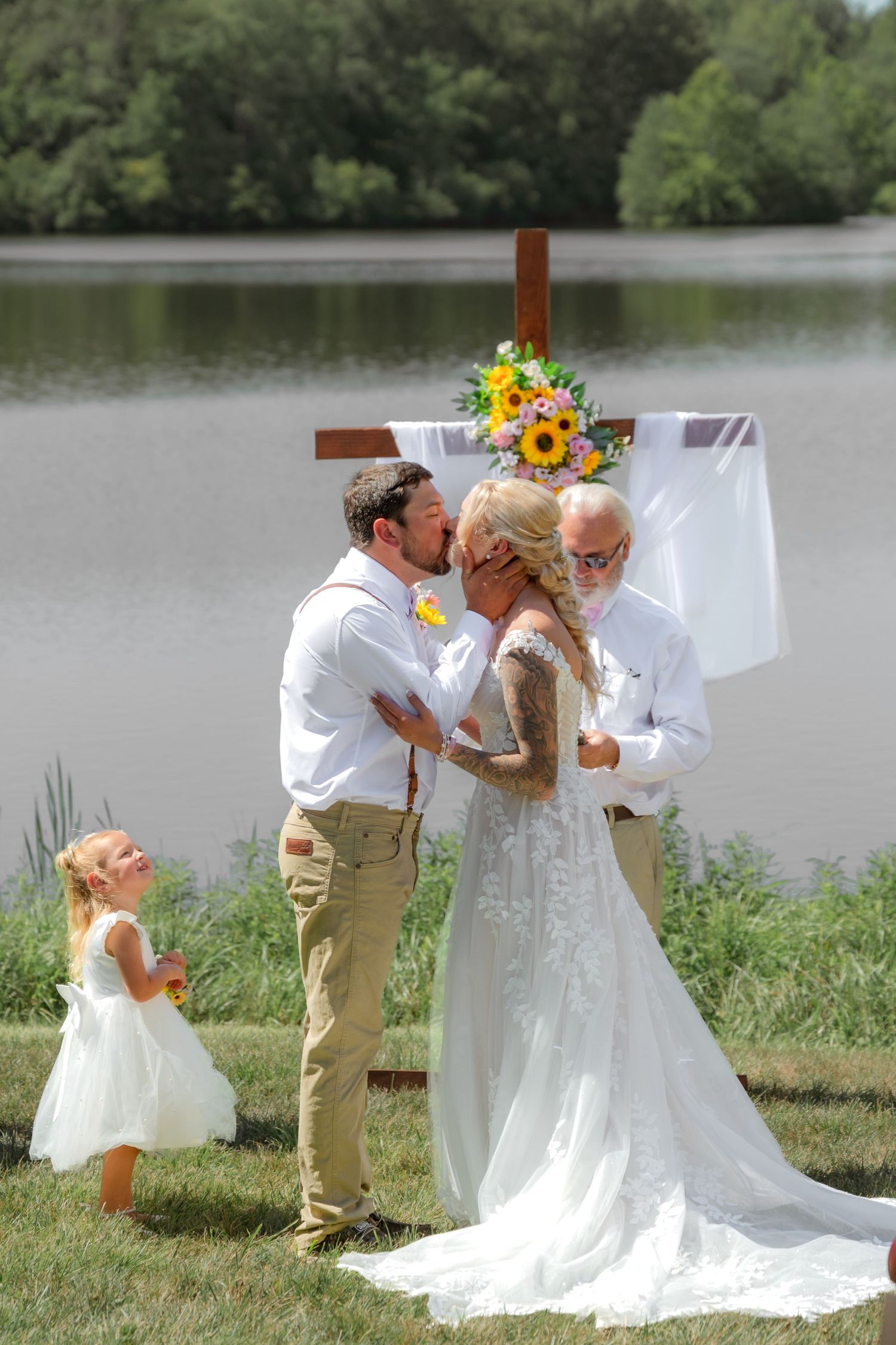 Outdoor wedding ceremony by a lake with couple kissing at wooden cross altar, flower girl, and sunflower bouquet.