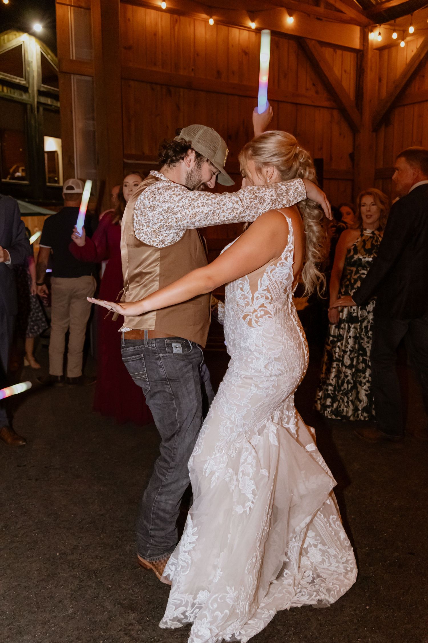Bride and groom share a romantic dance at rustic barn wedding reception with glowing sticks and guests in background.