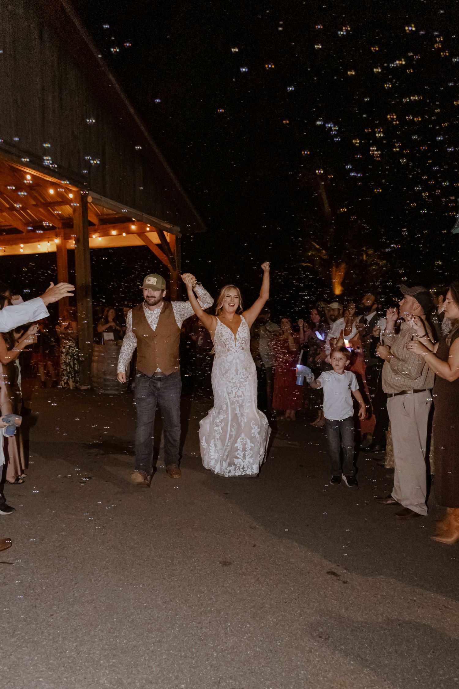 Newlywed couple celebrating wedding exit with bubbles, bride in lace gown, groom in vest and jeans, guests cheering.