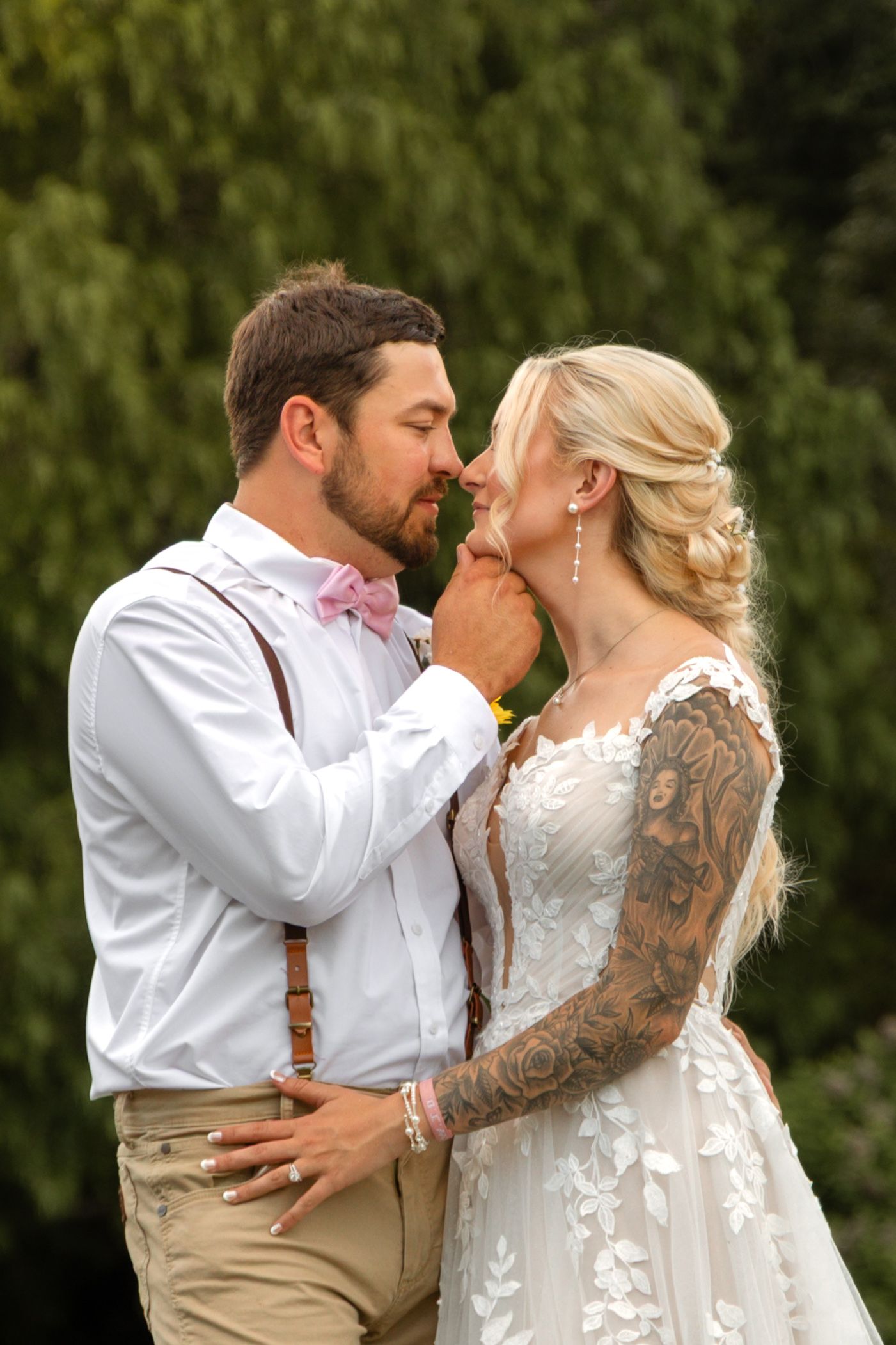 Bride and groom share a romantic moment outdoors, groom in suspenders and pink bow tie, bride in lace gown with tattoos.