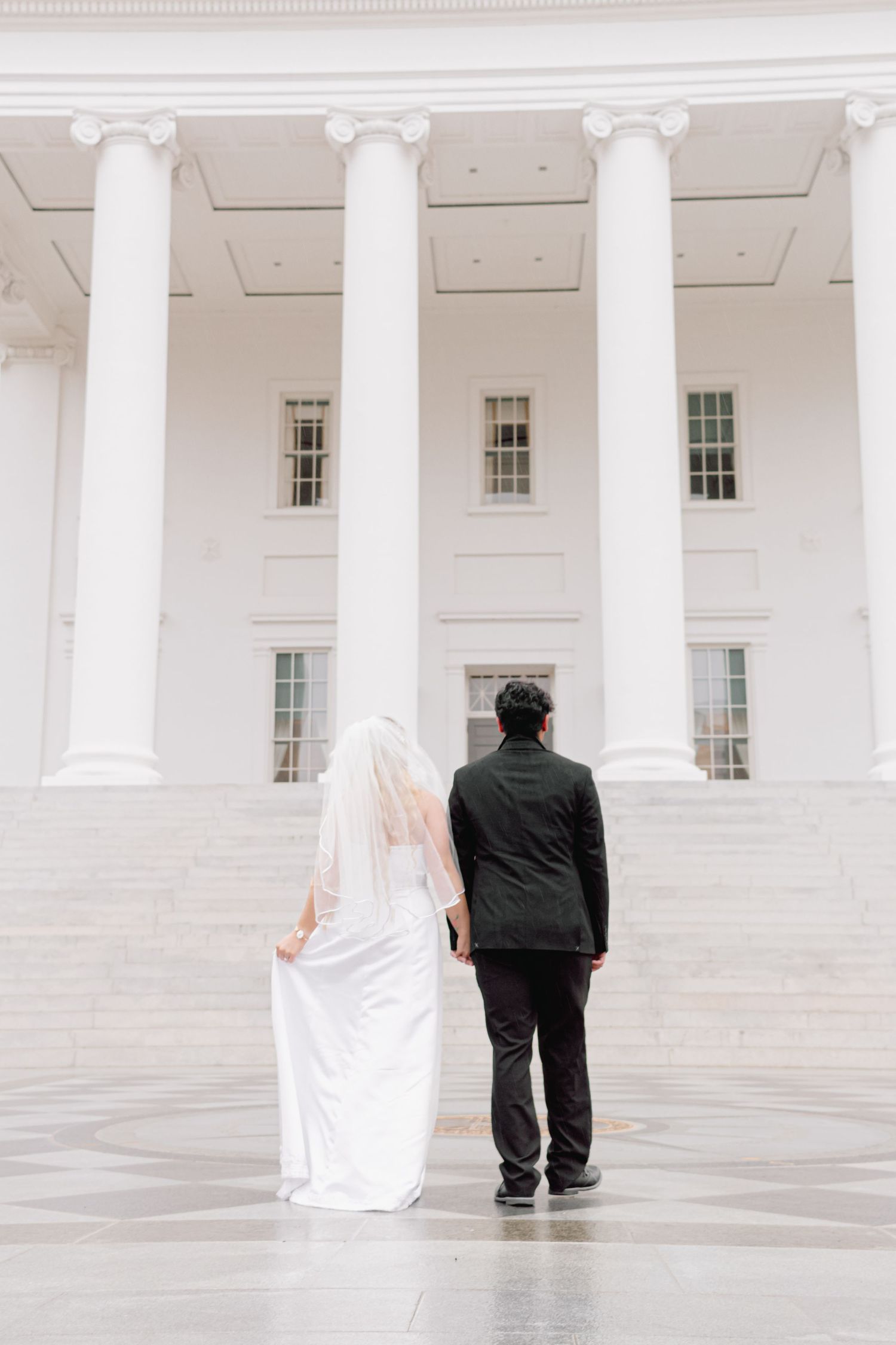 A couple stands together on white marble steps with grand columns in the background.
