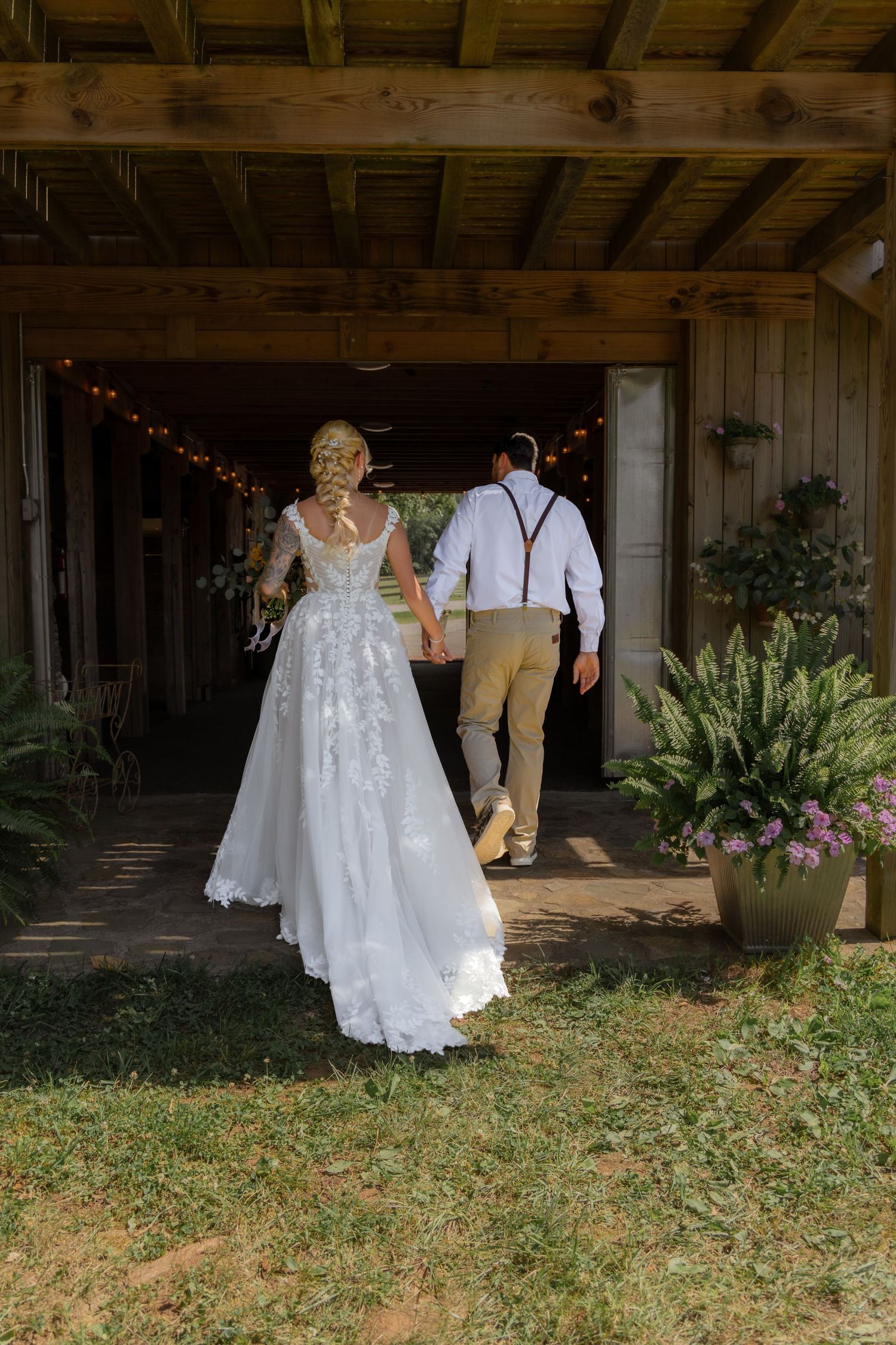 Bride in lace wedding gown walks hand-in-hand with groom in suspenders into rustic barn venue.