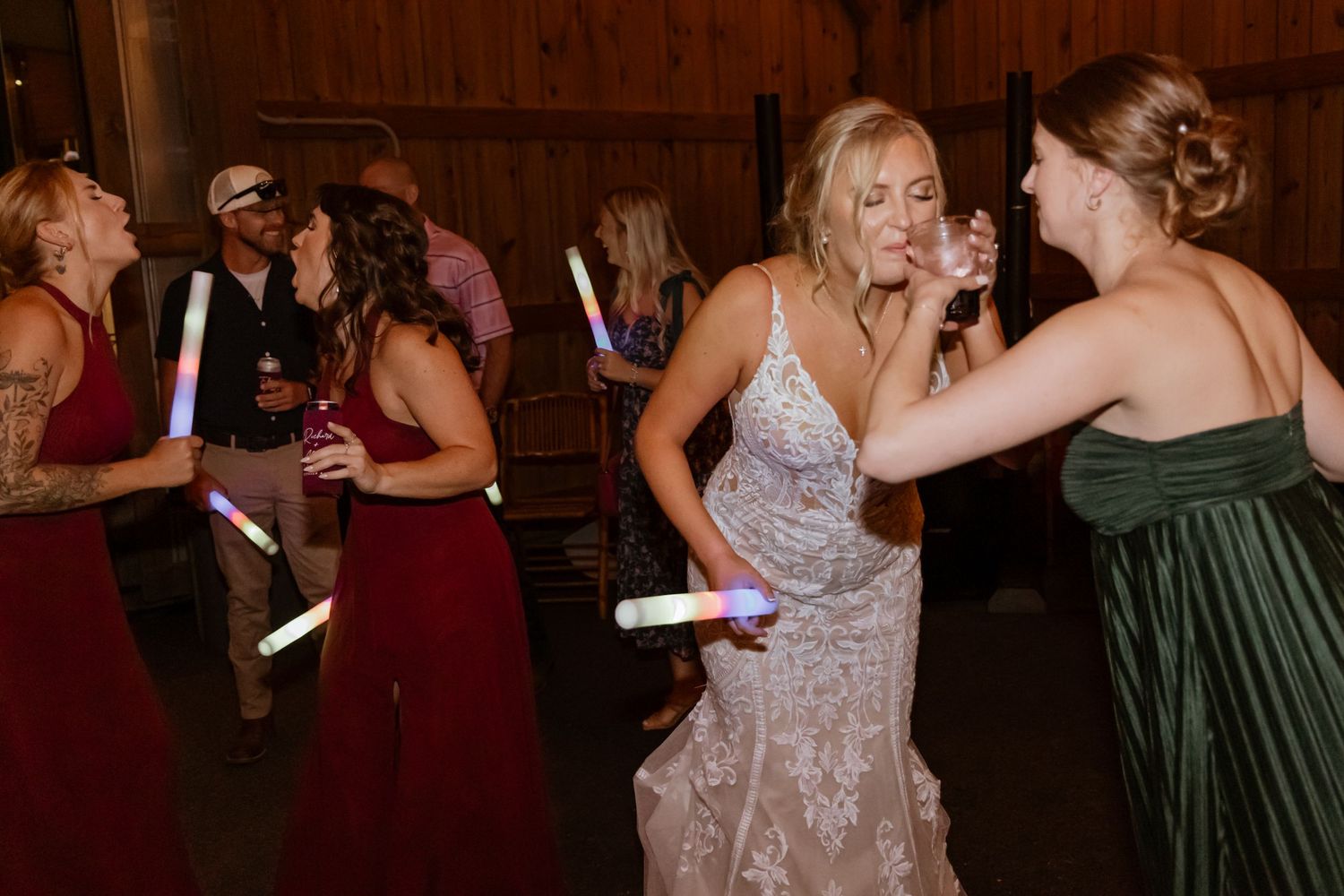 Bride dancing with bridesmaids in burgundy and green gowns holding glow sticks at a wedding reception.