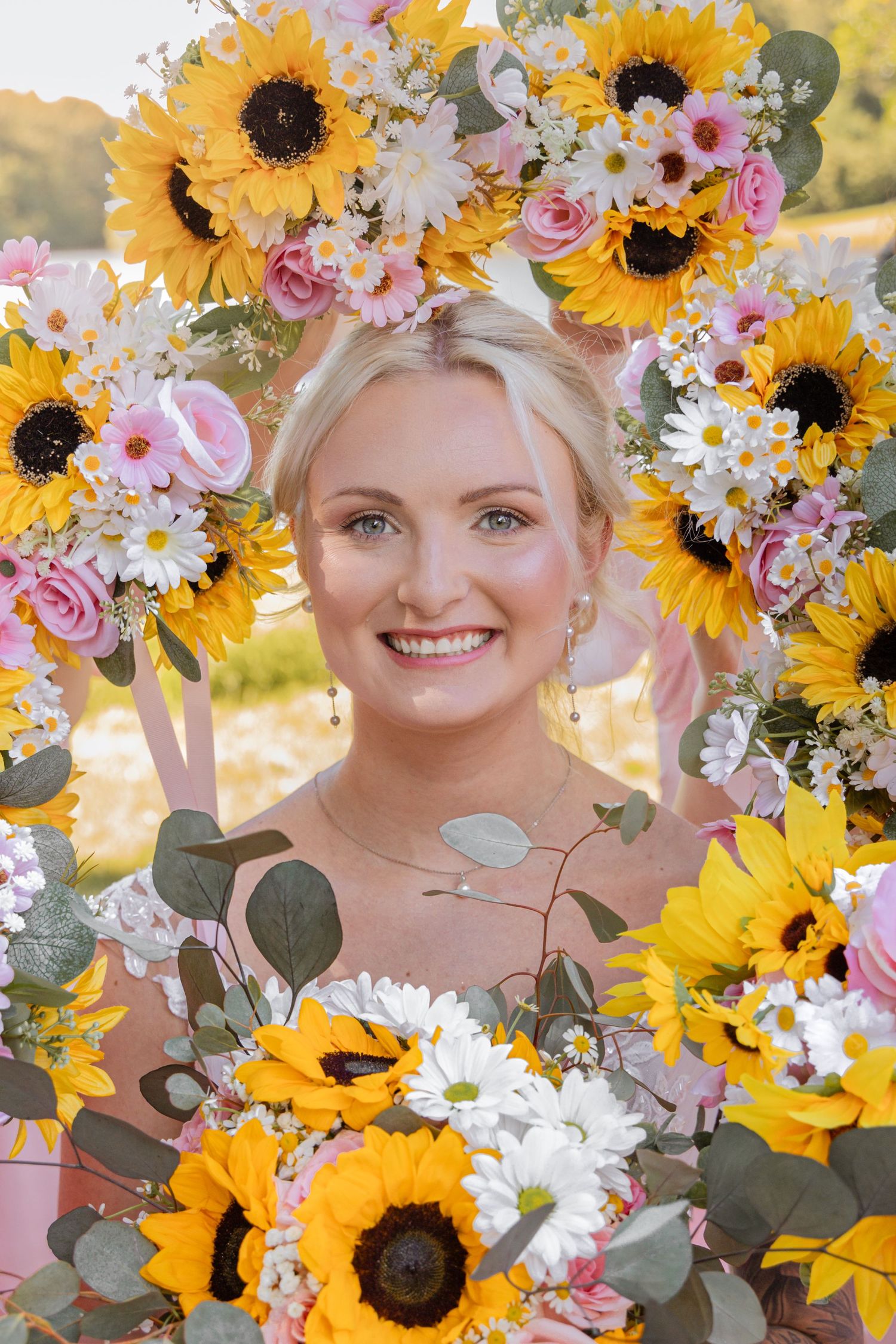 Smiling blonde woman framed by sunflowers, pink roses, and white daisies in a floral arrangement outdoors.