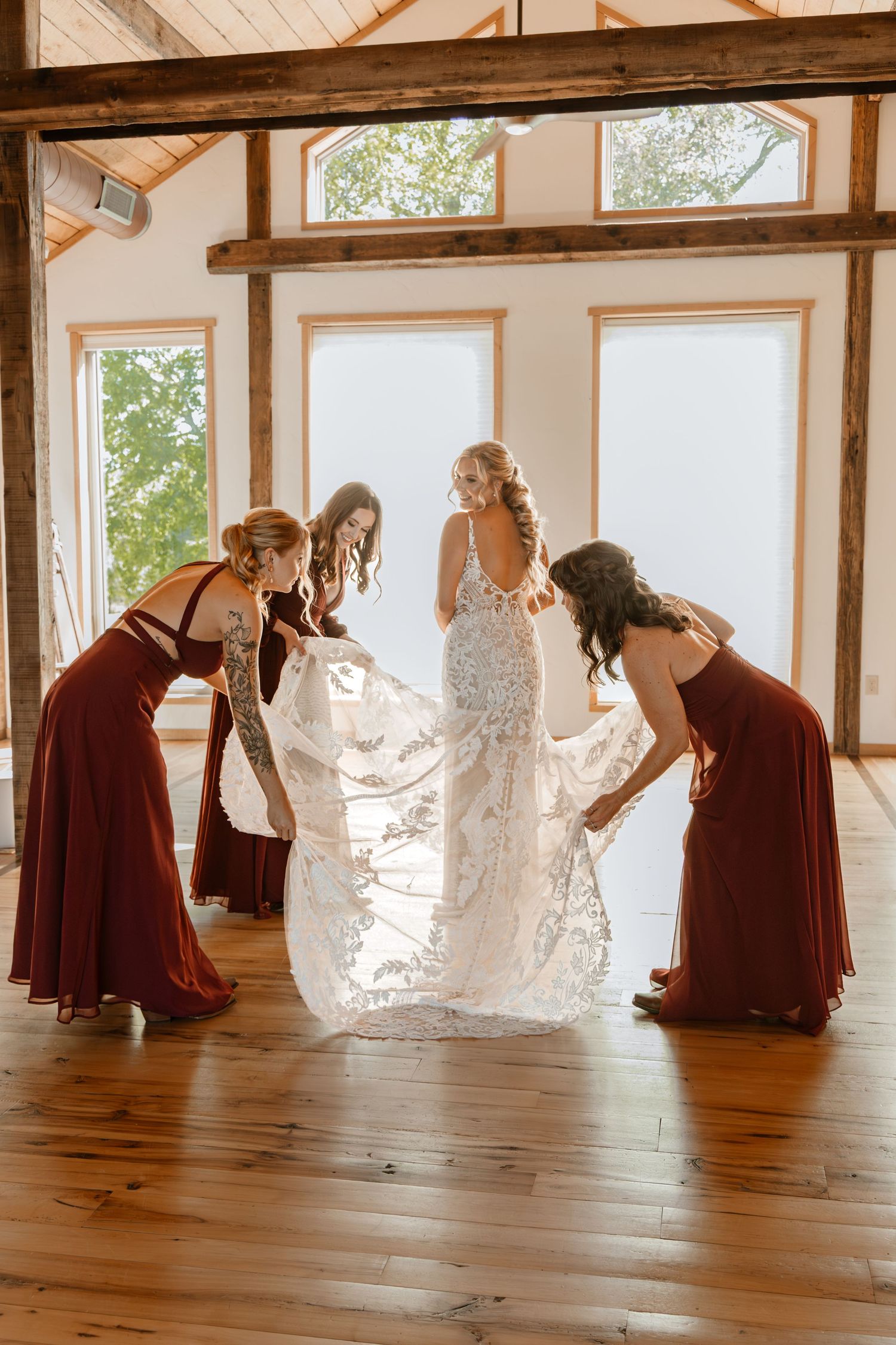 Bridesmaids in burgundy dresses help arrange bride's train in sunlit venue.