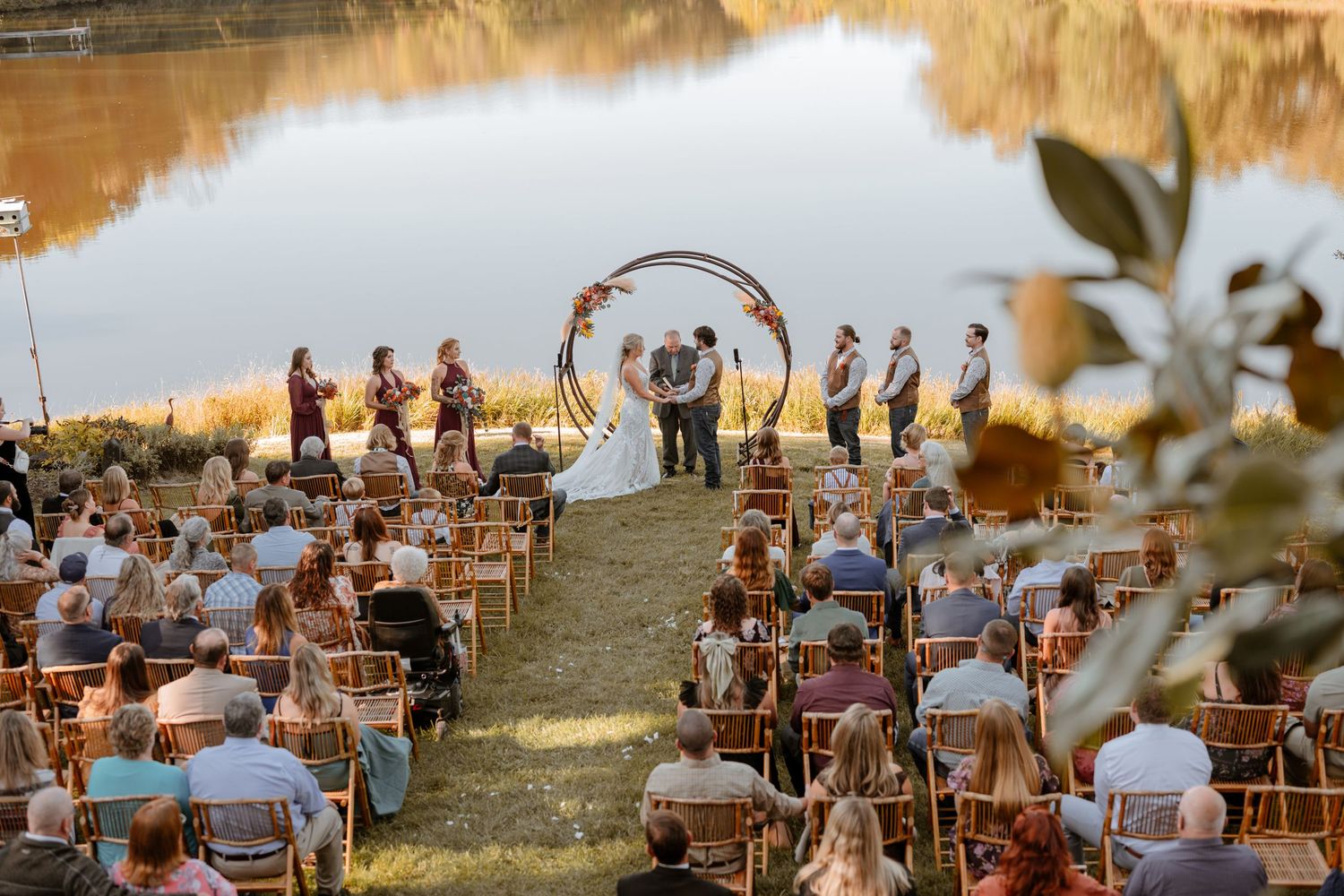 Panoramic view of lakeside wedding ceremony setup with wooden chairs and natural fall scenery.