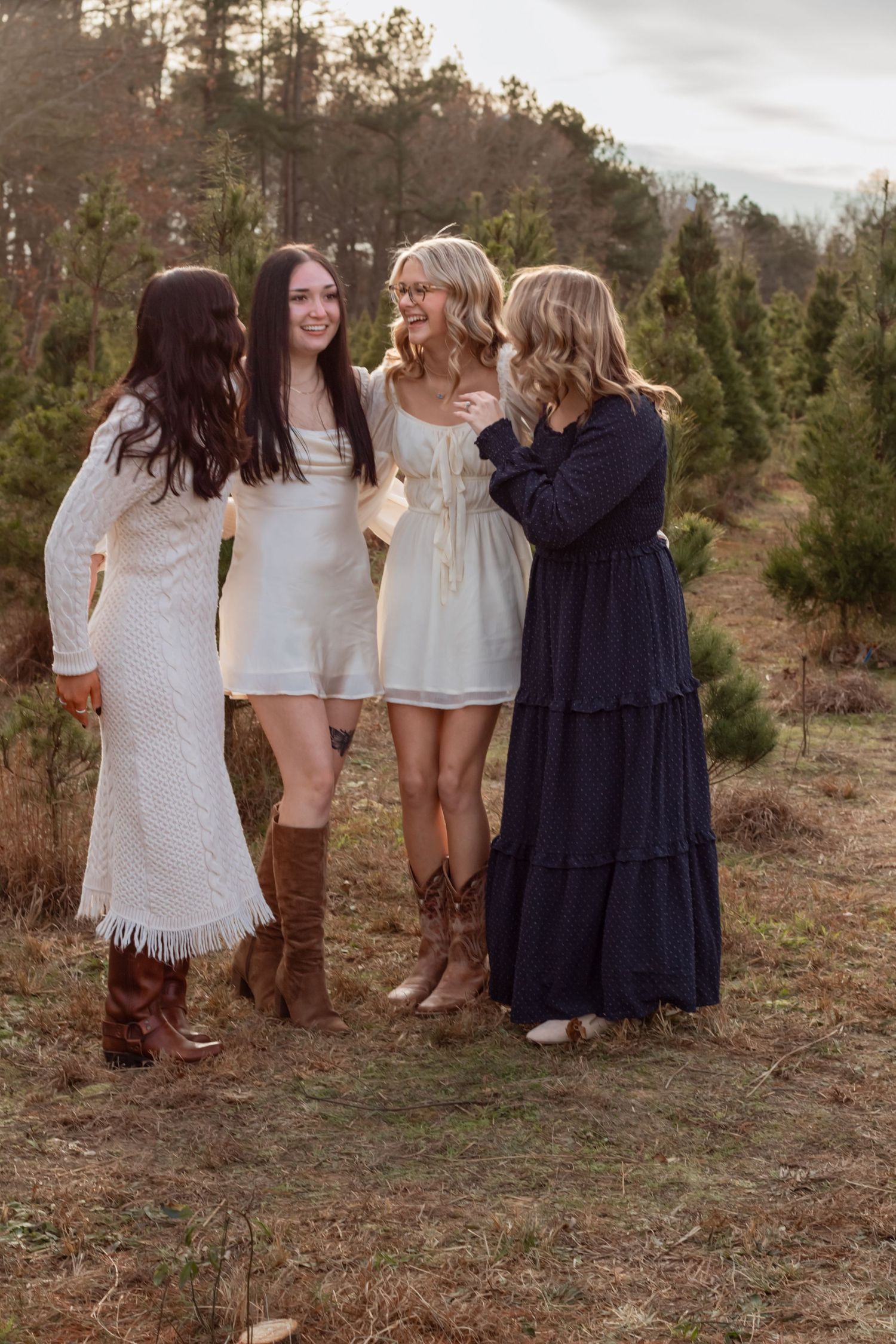 Four women in coordinated white and navy dresses laugh together at a Christmas tree farm.
