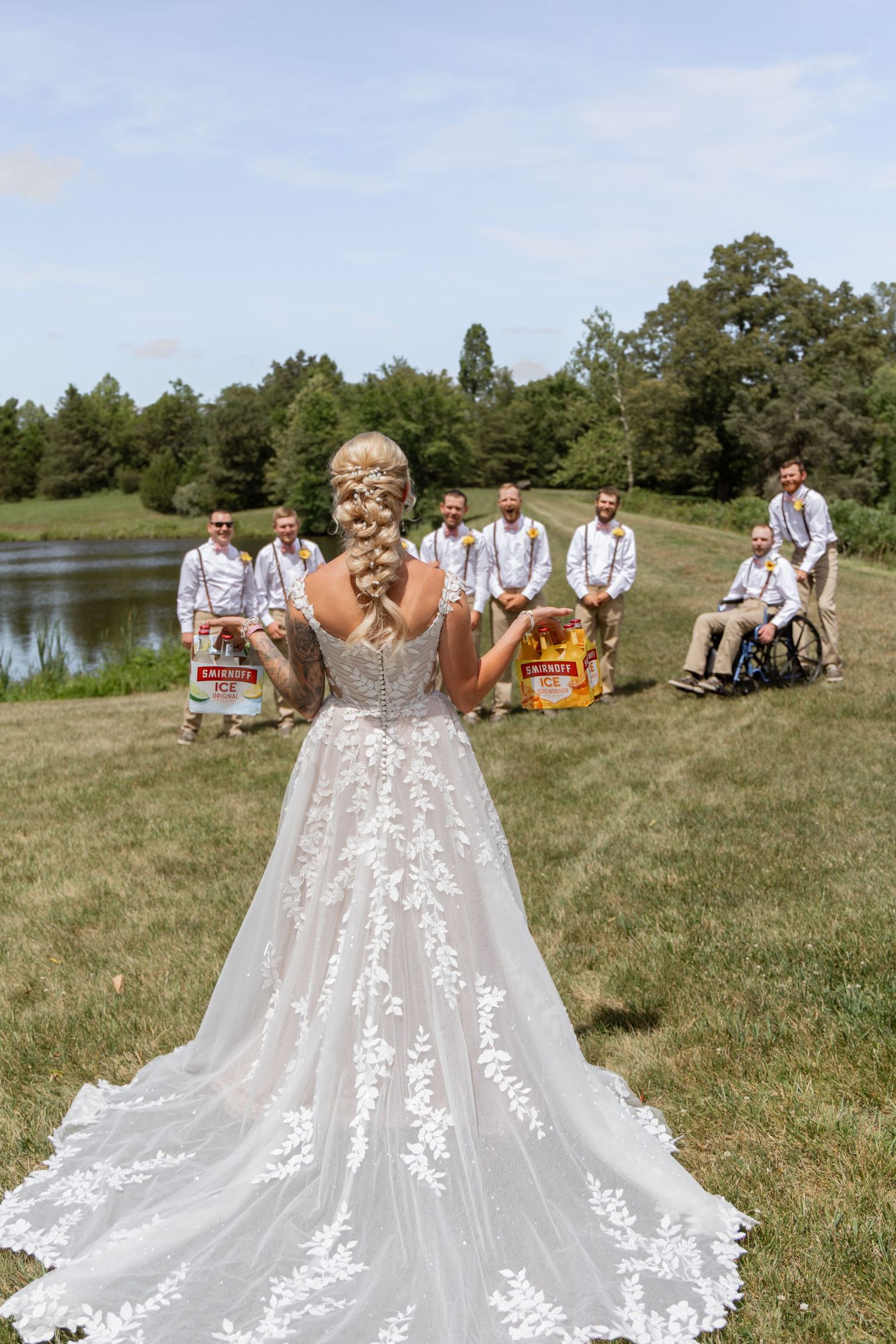 Bride in lace wedding gown faces groomsmen holding Busch Light cases by a pond.
