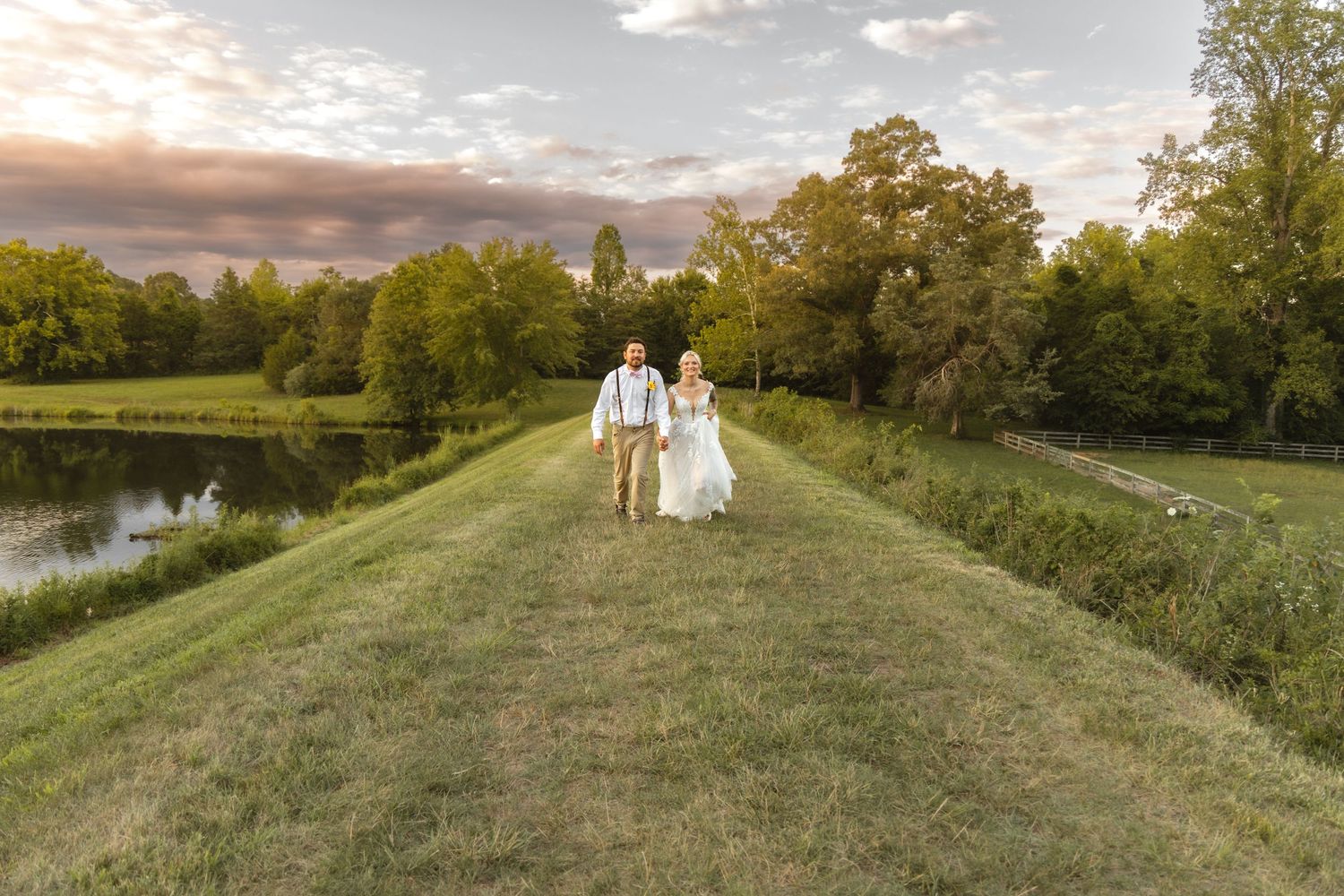 Bride and groom walk hand-in-hand along a grassy path at sunset near a pond.