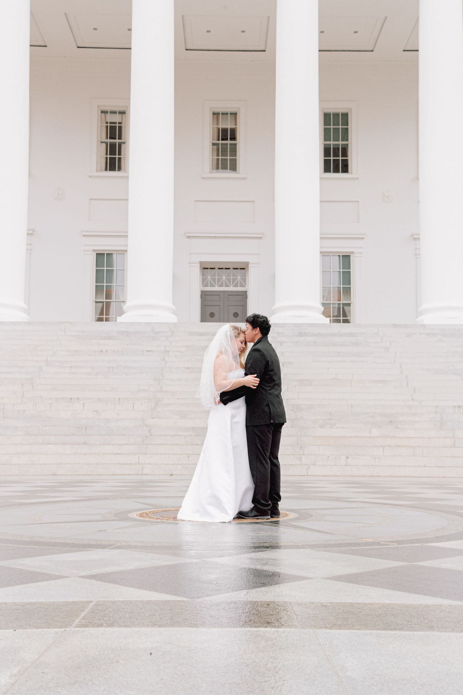 A romantic wedding couple shares their first kiss on the steps of a grand white columned building.