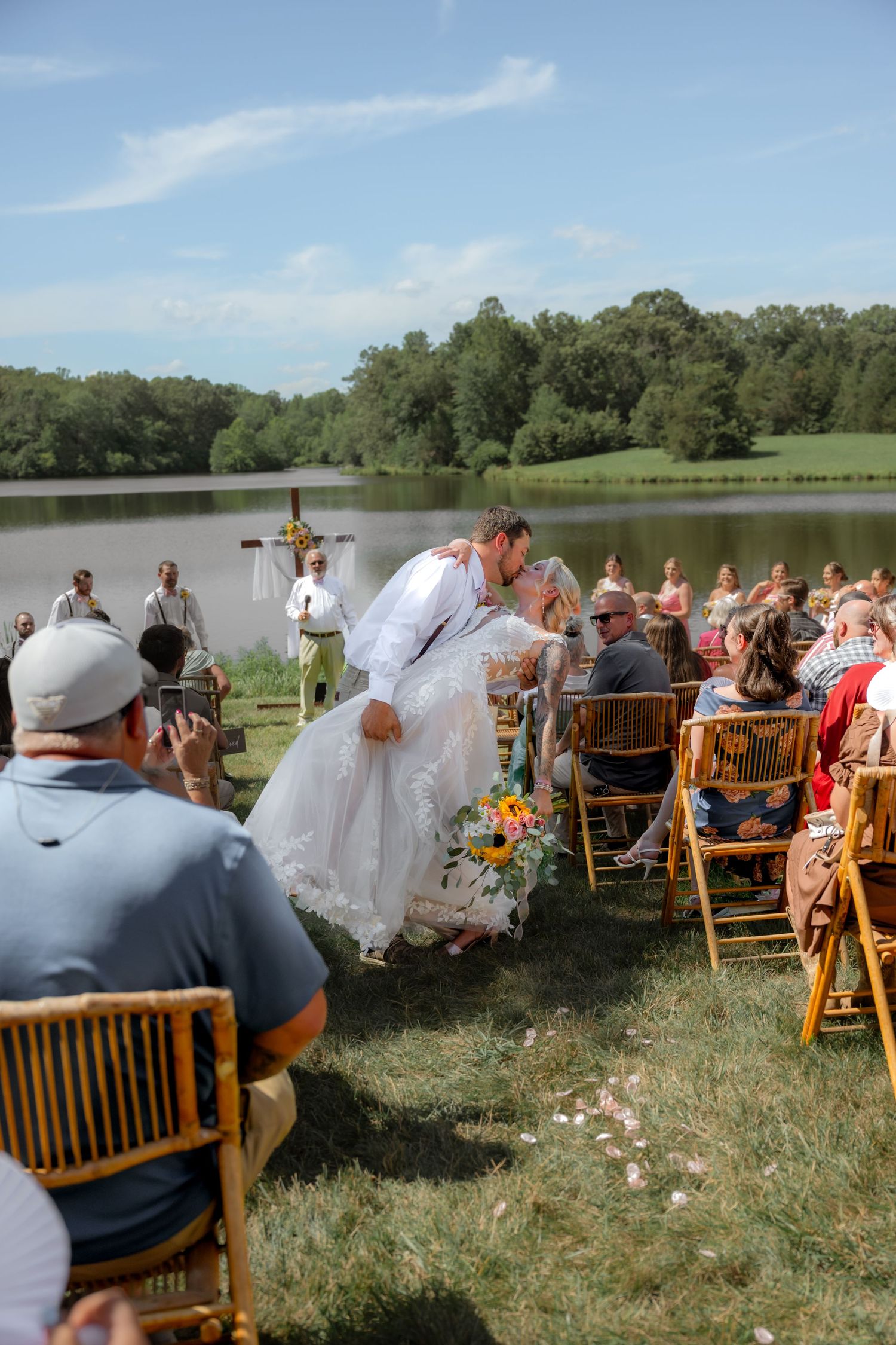 Outdoor wedding ceremony by a lake, bride and groom sharing a kiss at the altar with guests seated on wooden chairs.