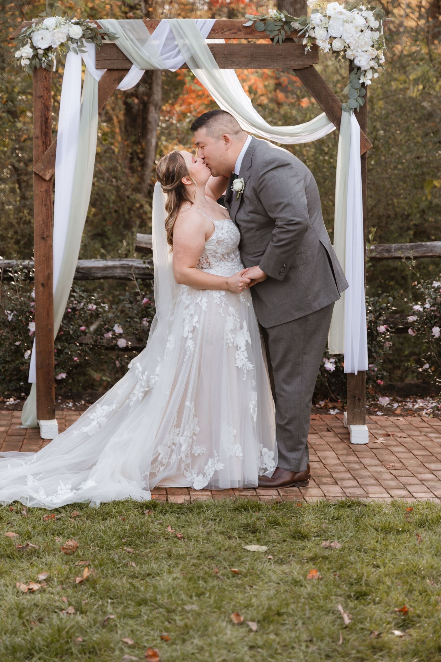 Wedding ceremony under white draped arch with brick pathway and autumn trees in background.