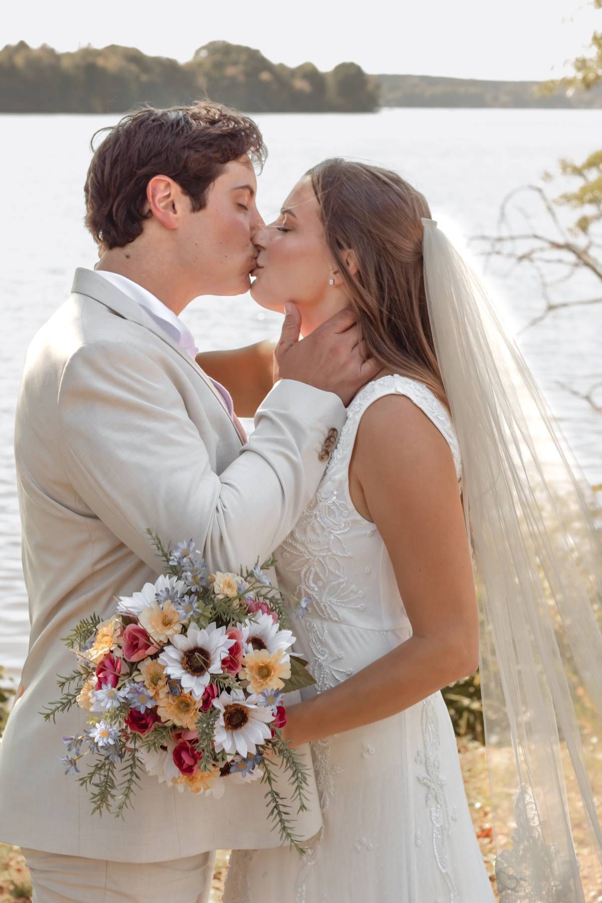 A romantic wedding kiss by the water at sunset with the bride holding a colorful floral bouquet.