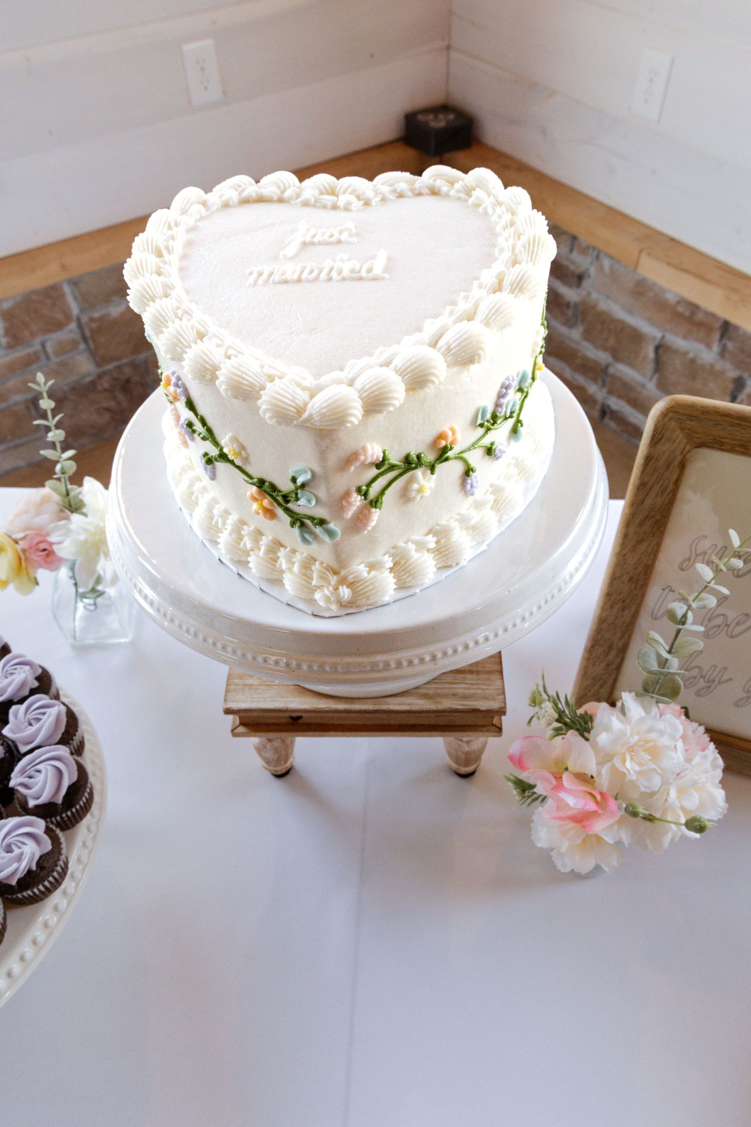 Heart-shaped white wedding cake with floral details and 'Just Married' topper on elegant cake stand.