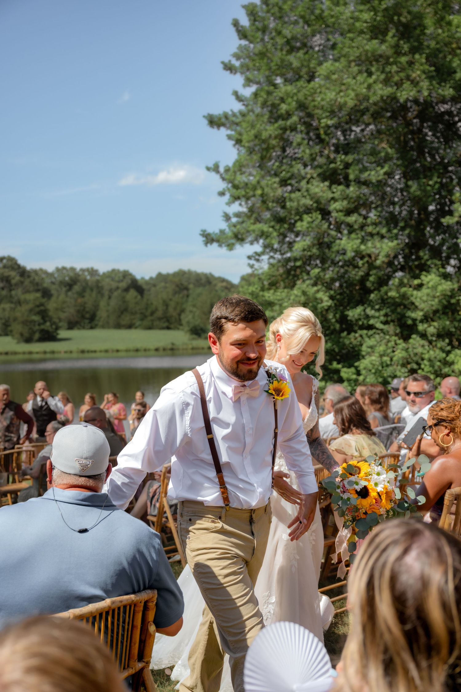 Bride and groom walk down the aisle at an outdoor wedding ceremony near a lake on a sunny day.