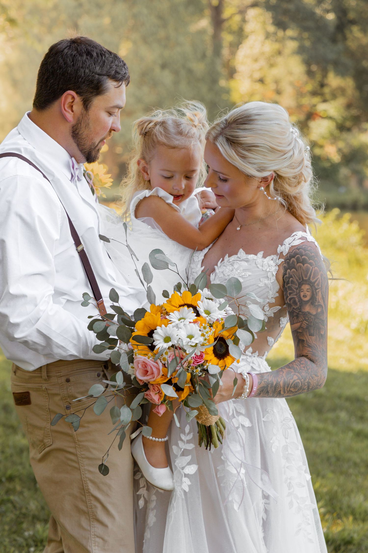 Bride and groom hold young daughter with sunflower wedding bouquet in golden outdoor setting.