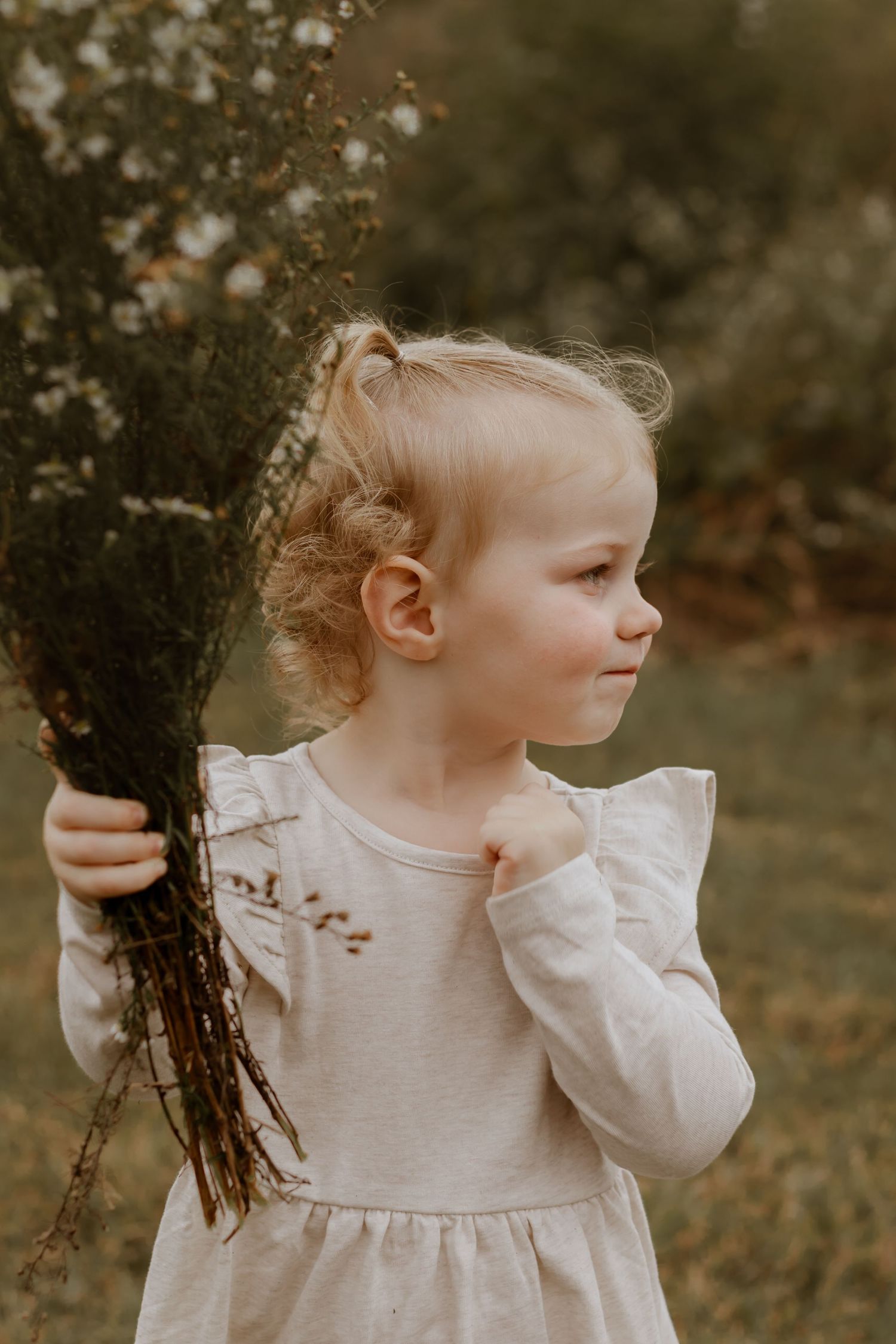 A young child in a white dress holds dried flowers while looking to the side in soft lighting.