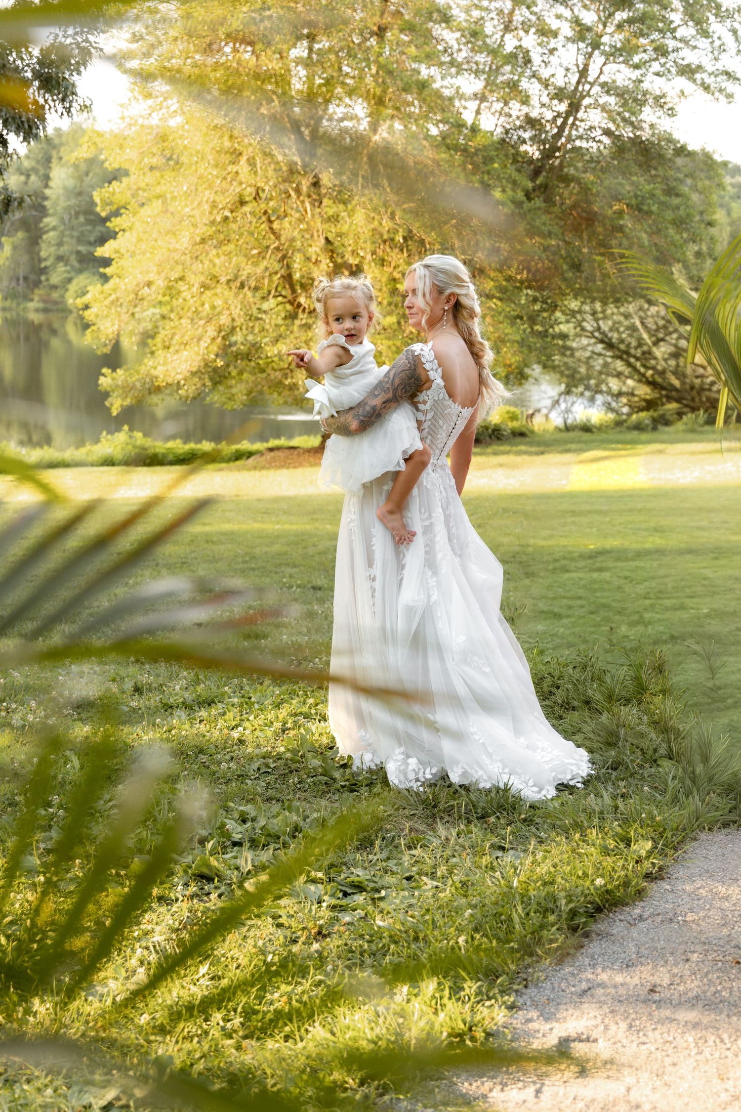 Bride in white lace gown holding a baby outdoors in golden sunlight among lush green trees.