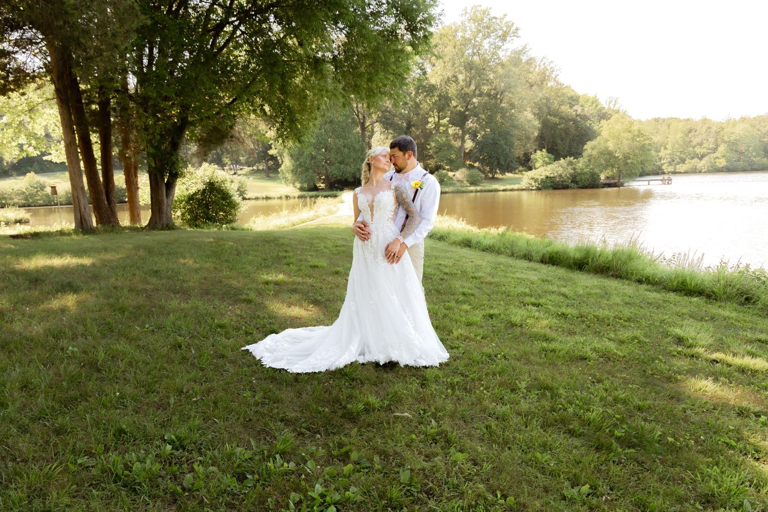 Bride and groom share a romantic kiss by a serene lakeside surrounded by lush green trees on their wedding day.