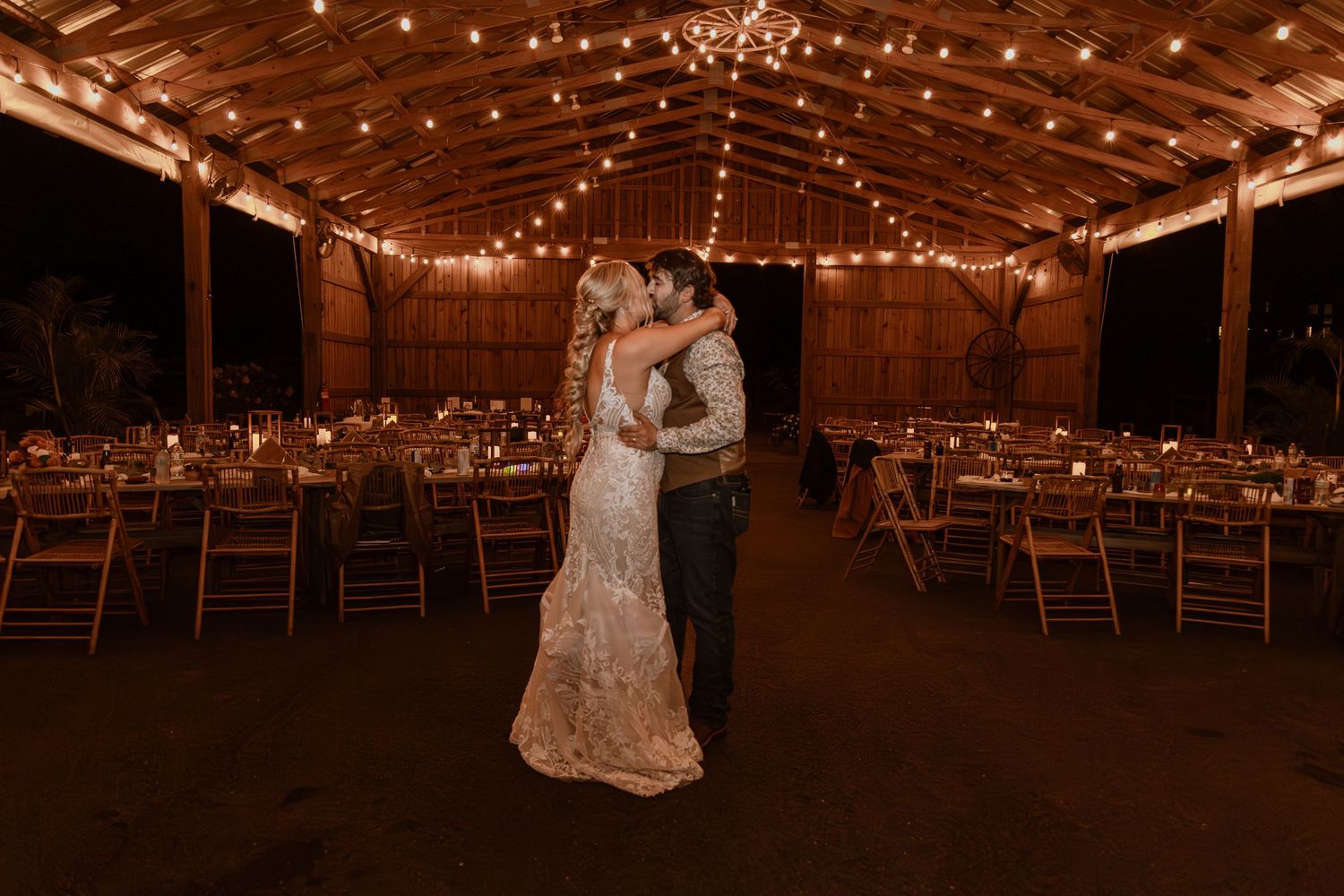 Bride and groom share first dance under warm string lights in a rustic barn wedding venue at night.