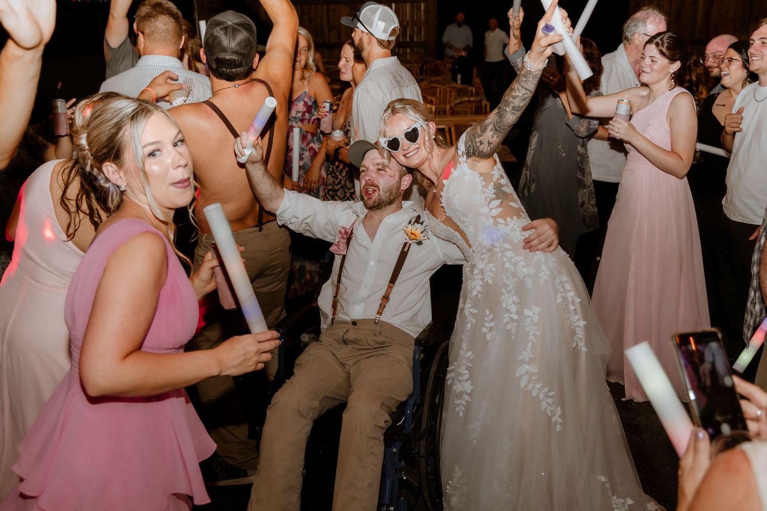 Bride and groom celebrating joyfully on dance floor with guests holding glow sticks at lively wedding reception.