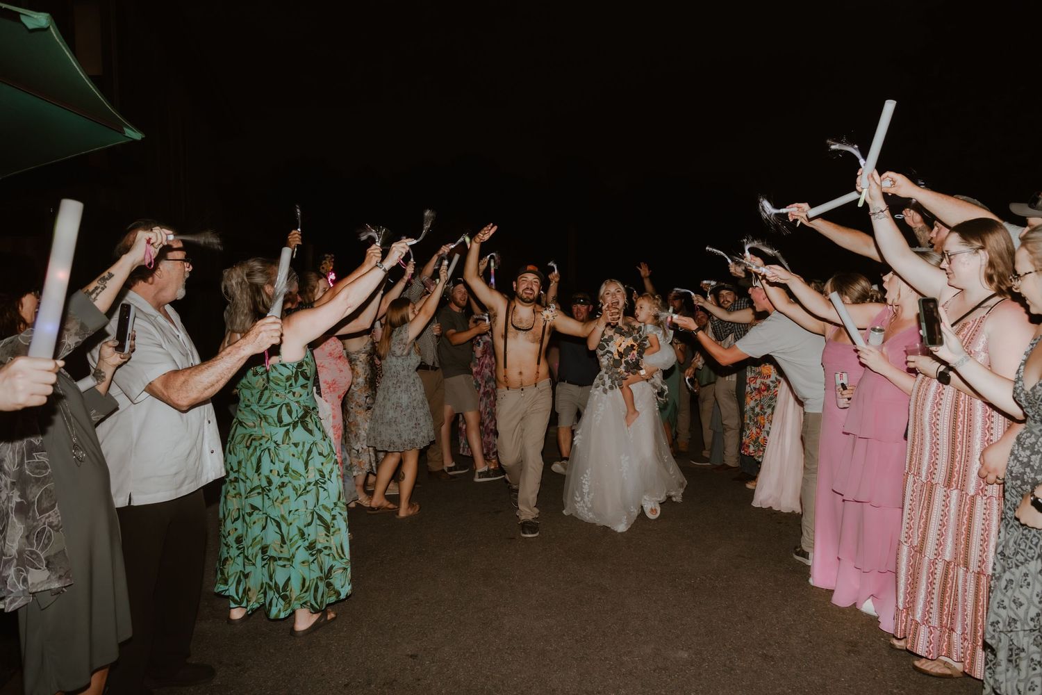 Wedding couple exits through a large crowd of guests holding glowsticks aloft at an outdoor evening celebration.