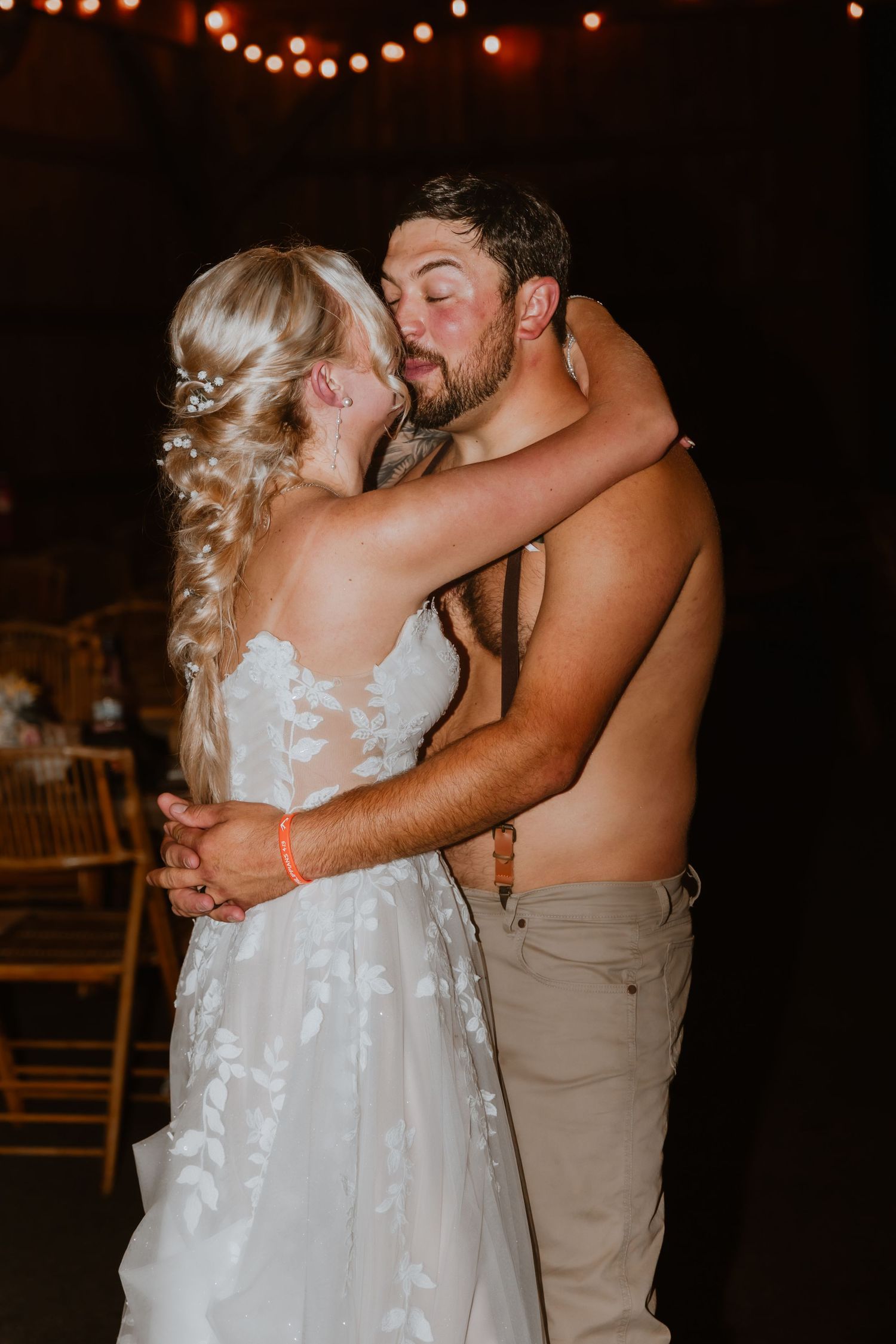 Bride in floral lace gown shares romantic first dance kiss with shirtless groom at rustic wedding reception.