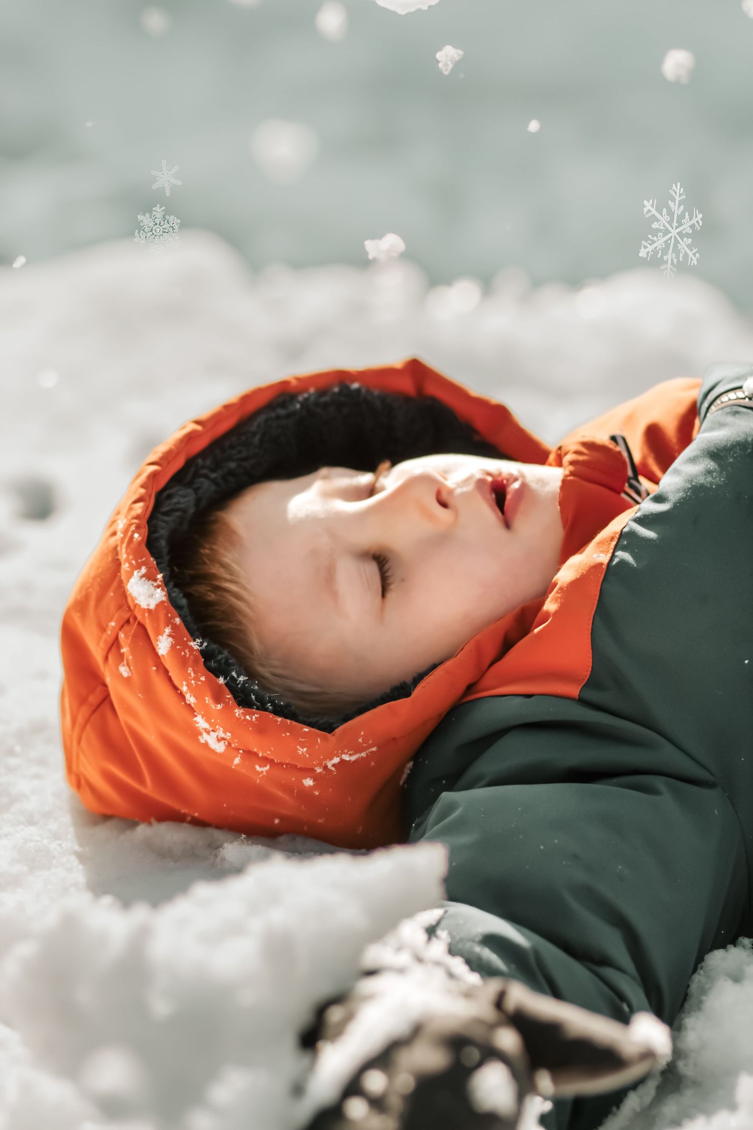 Person lying in snow wearing an orange hood and dark jacket with snowflakes falling.