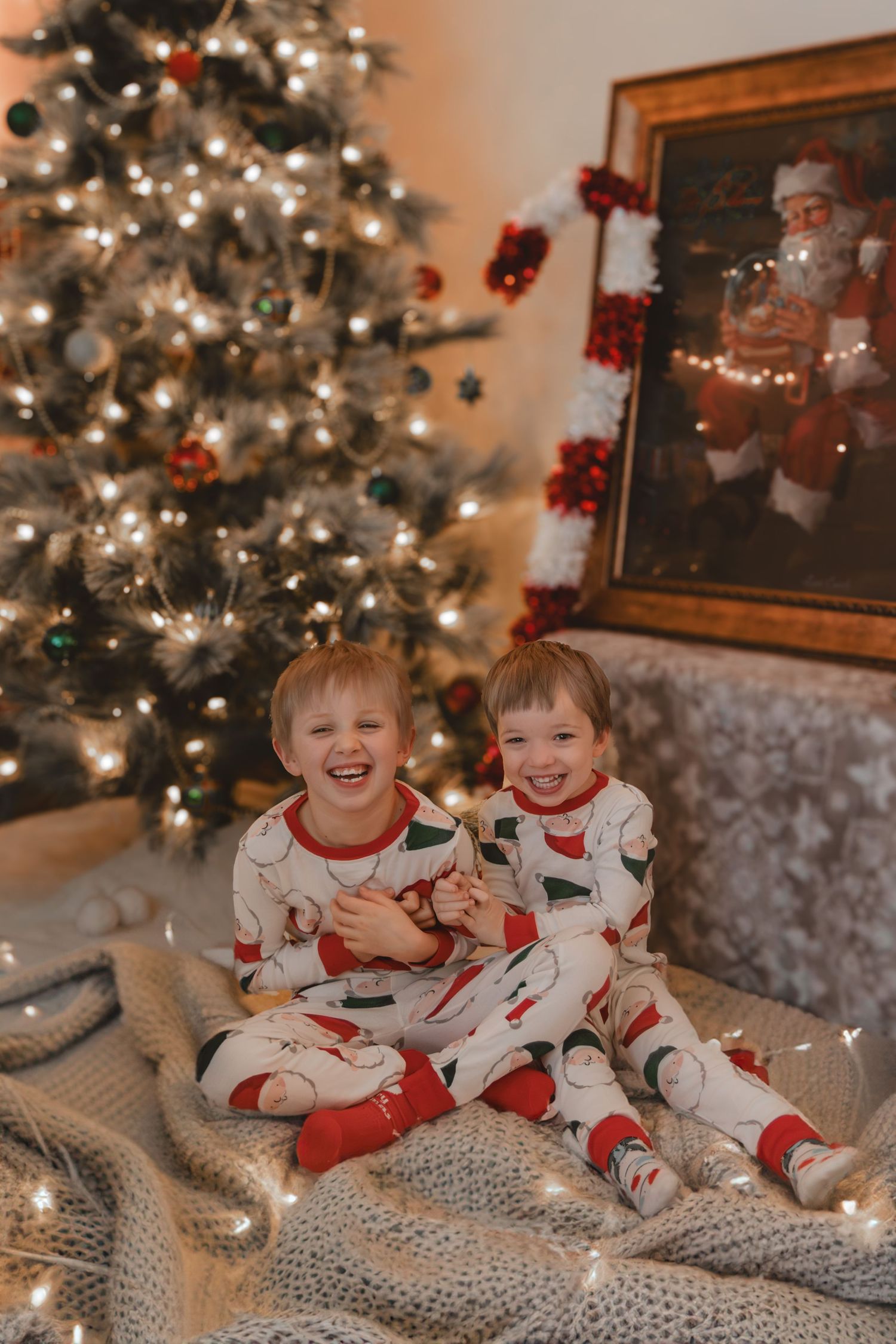Two children in matching Christmas pajamas laugh on blanket by decorated tree.