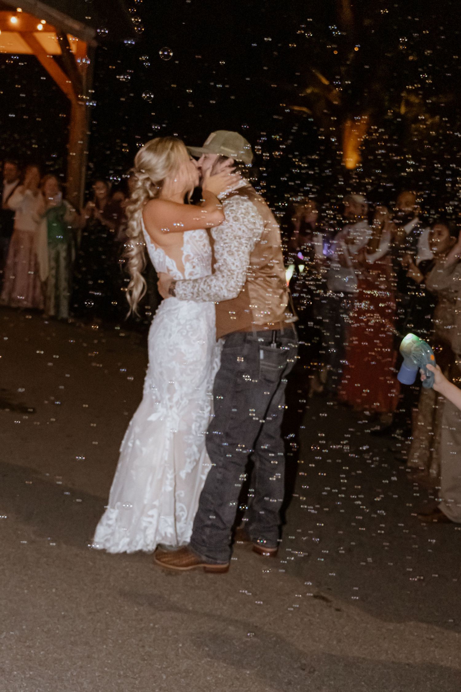 Newlywed couple sharing a romantic kiss surrounded by bubbles during their outdoor wedding reception at night.