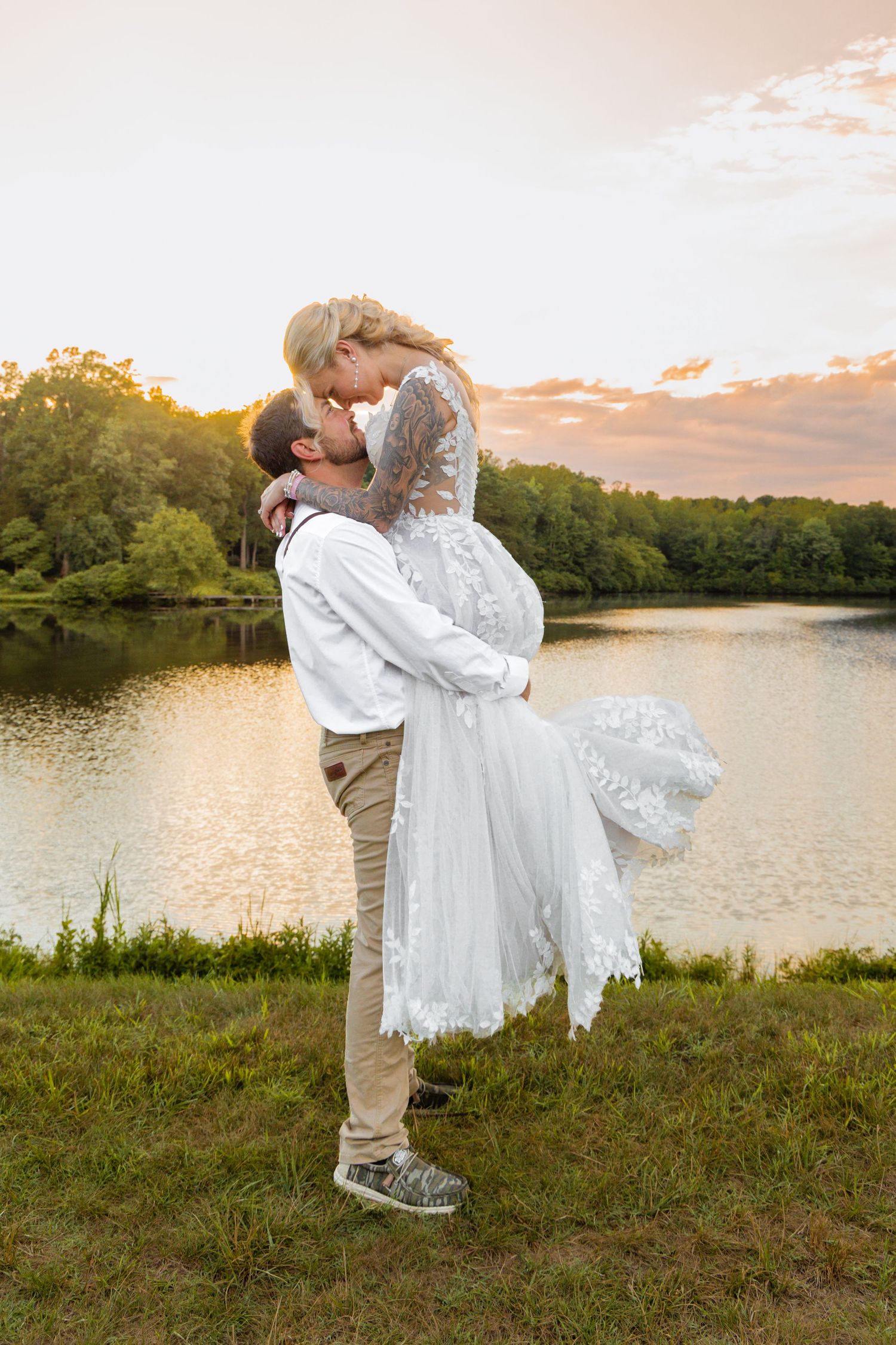 Groom lifts bride in white lace dress for a romantic kiss by a sunlit lake at sunset.