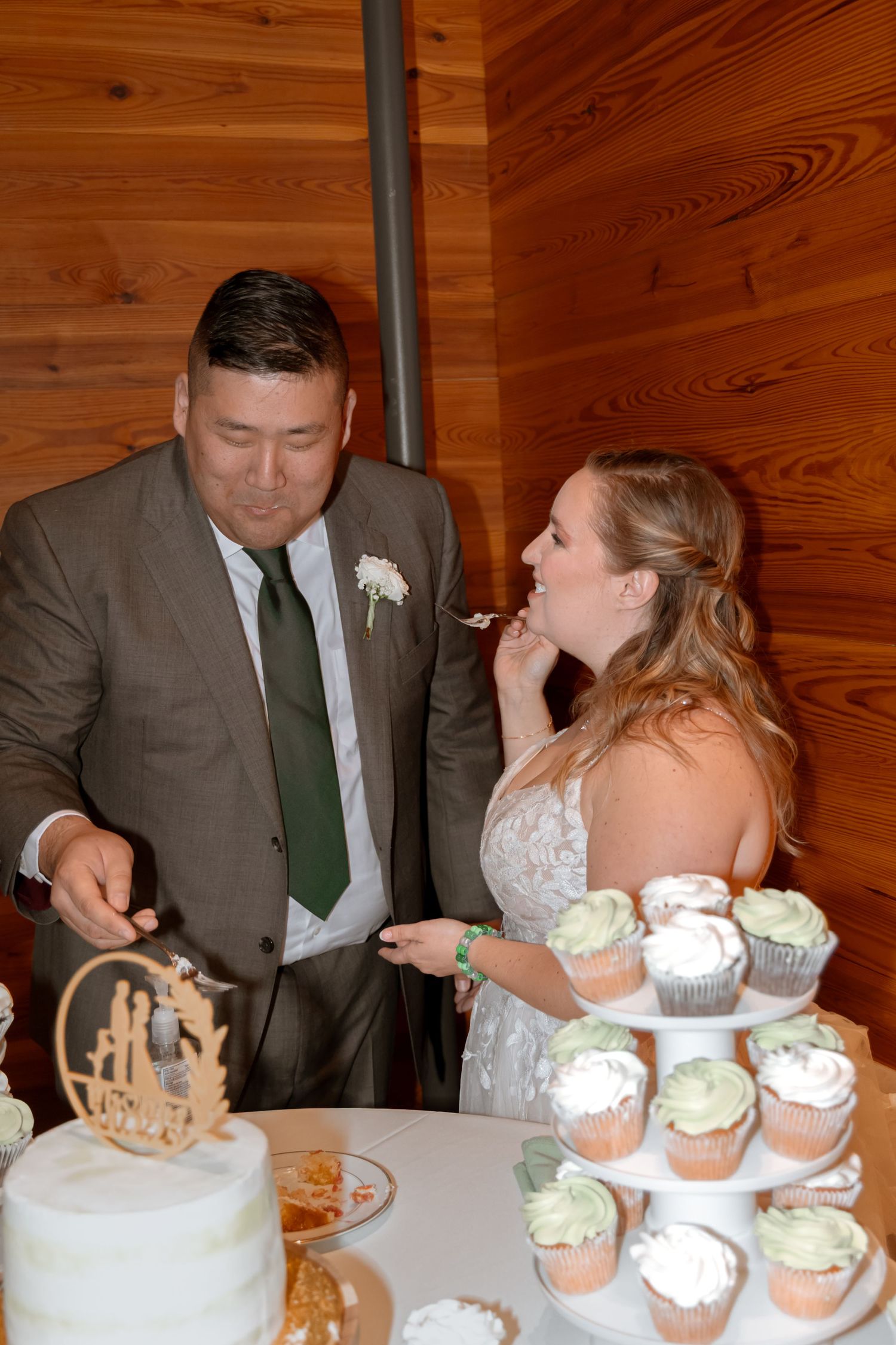 Wedding couple sharing cake and cupcakes at rustic wooden reception table.