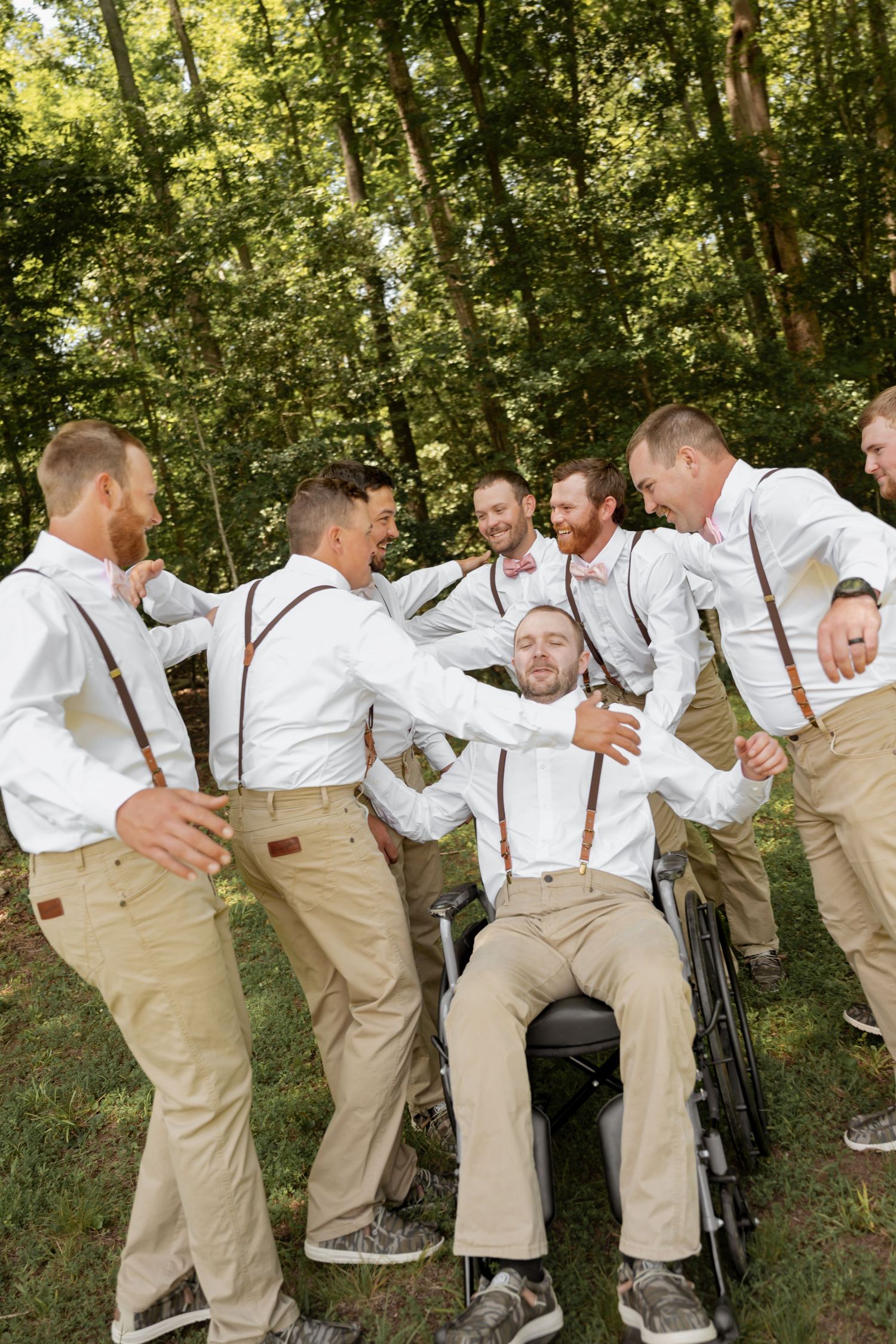 Groomsmen in white shirts and suspenders celebrate with groom in wheelchair outdoors at rustic wedding.