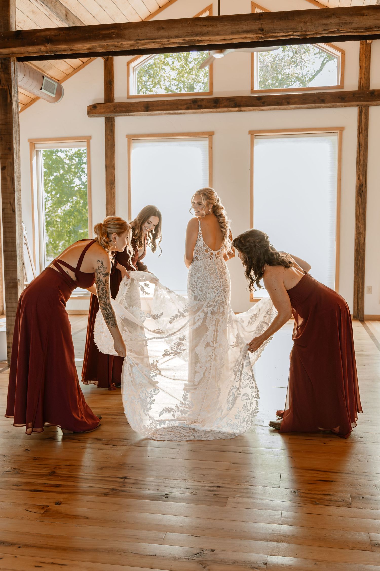 Wedding party members help adjust a bride's dress in a sunlit rustic venue.