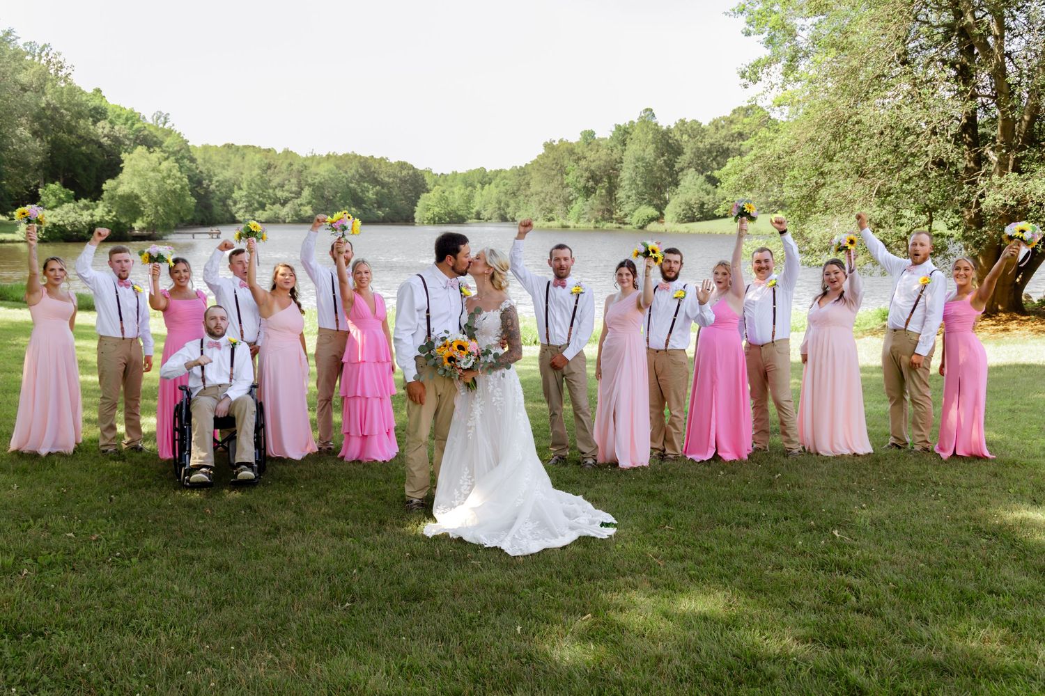 Wedding party poses outdoors by a lake, bridesmaids in pink dresses, groomsmen in khaki, as bride and groom share a kiss.